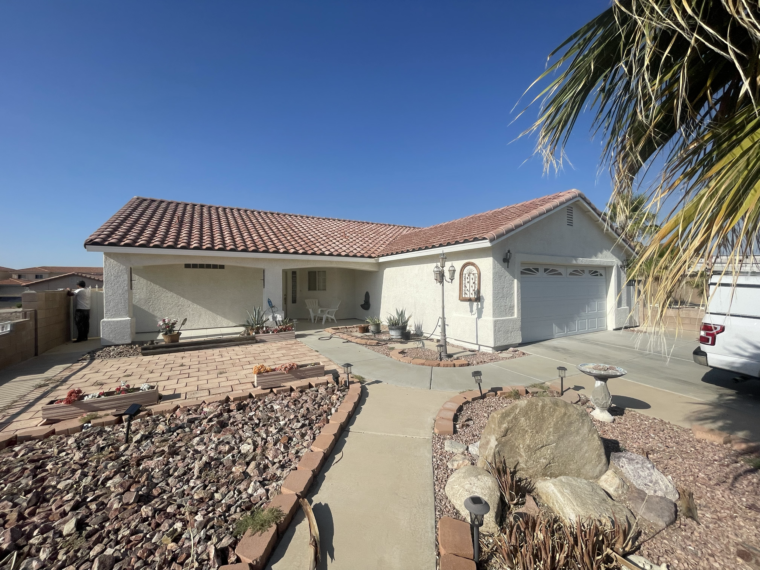 Front view of a single-story house with a red-tile roof, white exterior walls, a garage, and a landscaped yard with rocks, plants, and decorative elements under a clear blue sky.