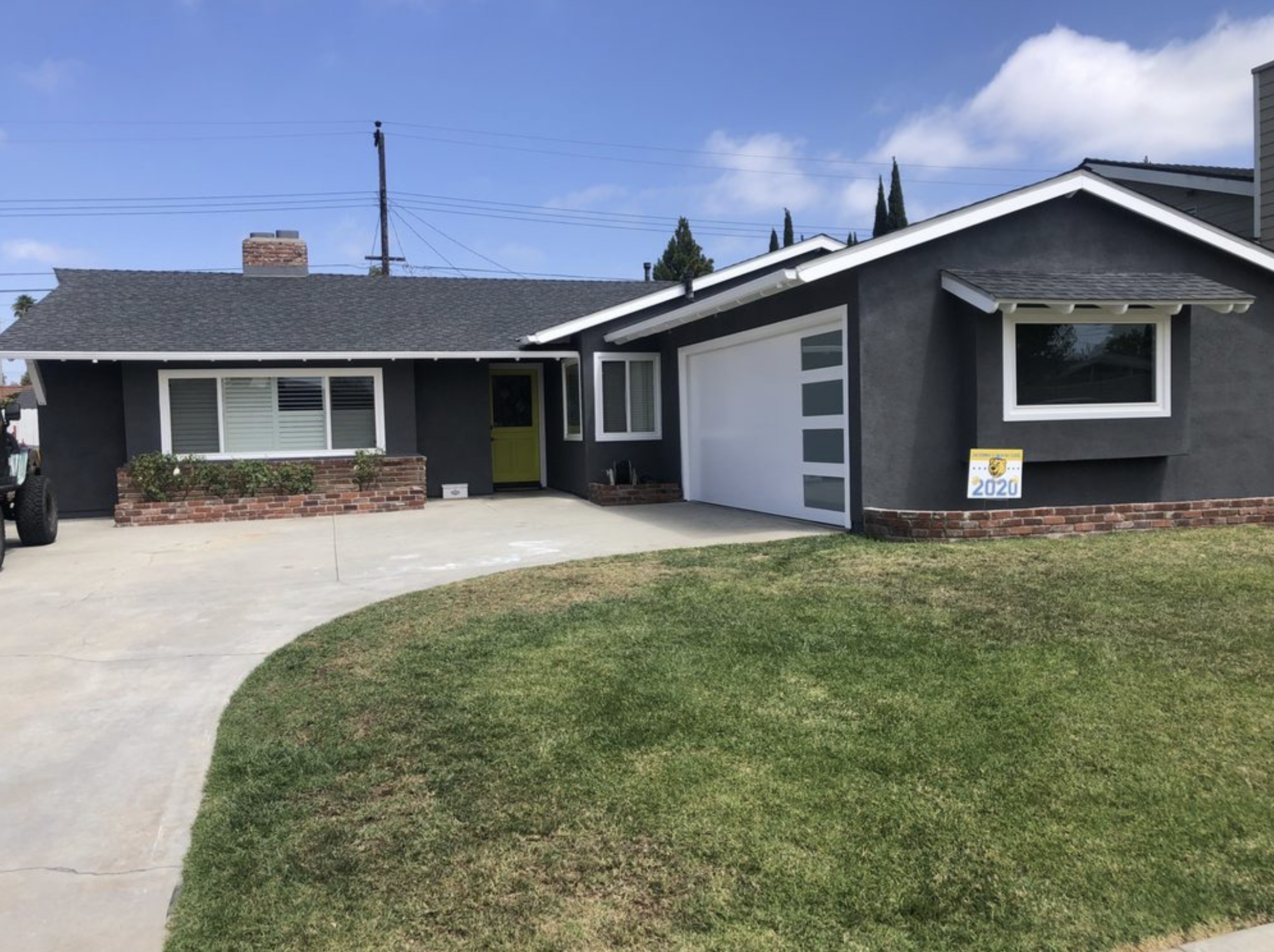 Front view of a single-story black house with white trim, a yellow front door, a white garage door with horizontal windows, landscaped brick flower beds, and a green lawn. A porch sign indicating the year 2020 is next to the right window.