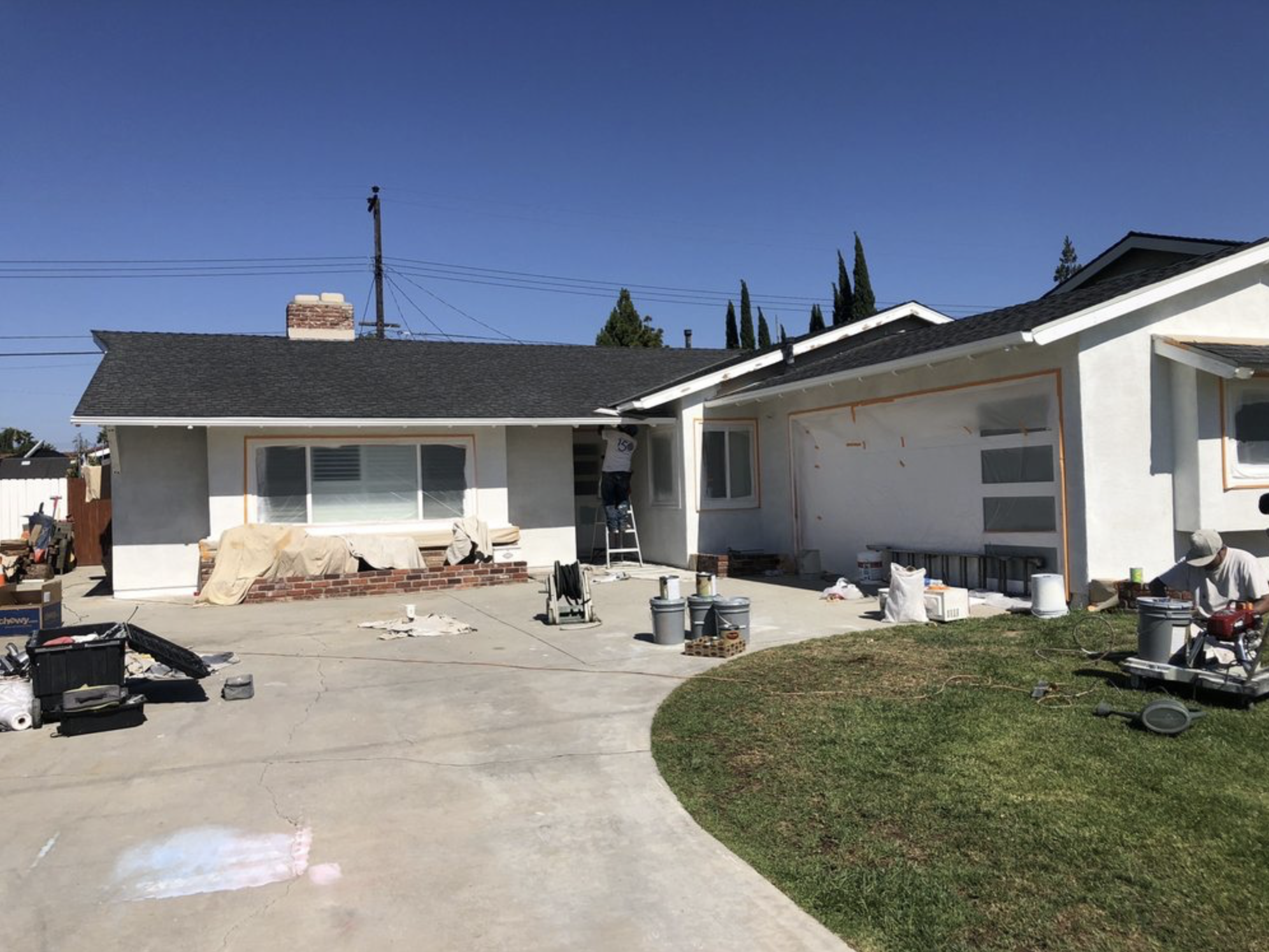 A house is undergoing exterior renovation, with workers painting and doing construction work. The house has white walls, black roof, and brick accents. Construction tools and materials are scattered in the driveway and yard.
