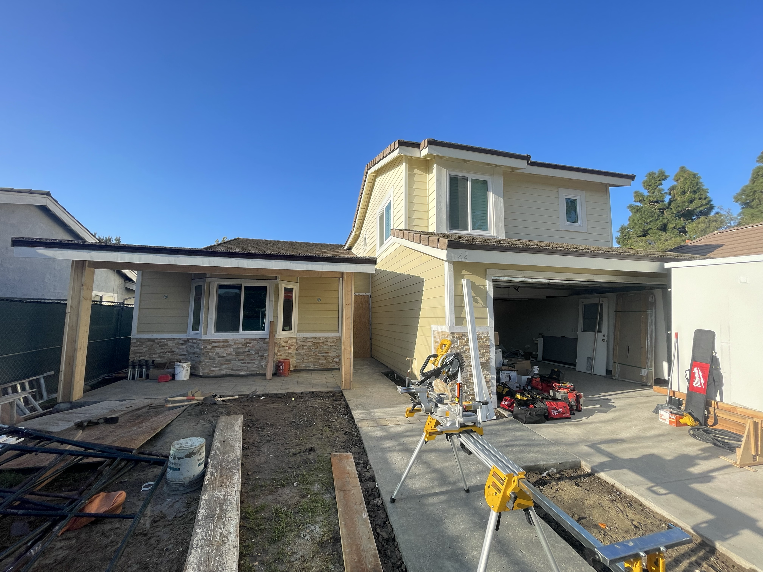 A two-story house under construction with a yellow exterior, white trim, and stone accents on the lower portion. The front yard is unfinished with construction tools and equipment, including a yellow saw on a tripod, scattered around. The garage is o