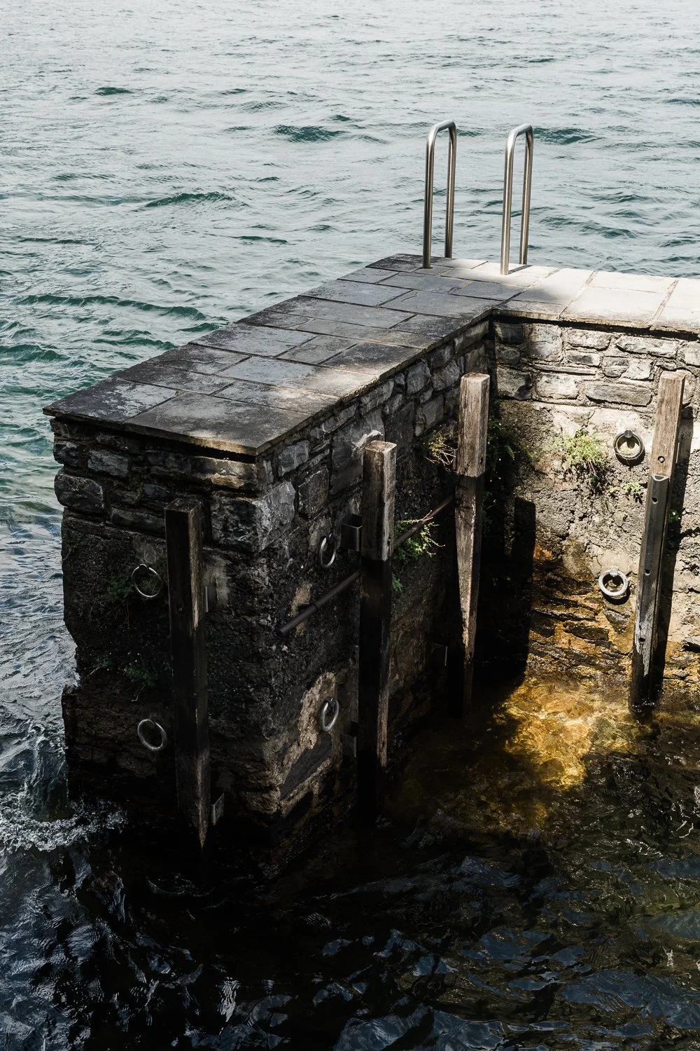 Ancient stone pier and swimming dock with iron mooring rings and metal ladder extending into the waves of Lake Como, Varenna, Italy — Italy travel photographer Anna Hudson
