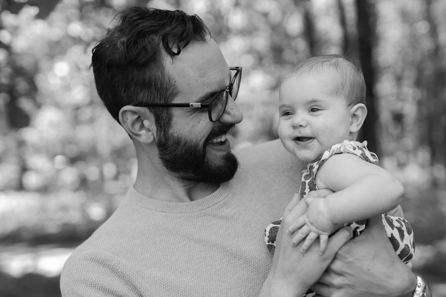 Black and white candid portrait of a father laughing with his smiling baby daughter among bokeh trees at Forest Park, St. Louis, Missouri — family photographer Anna Hudson