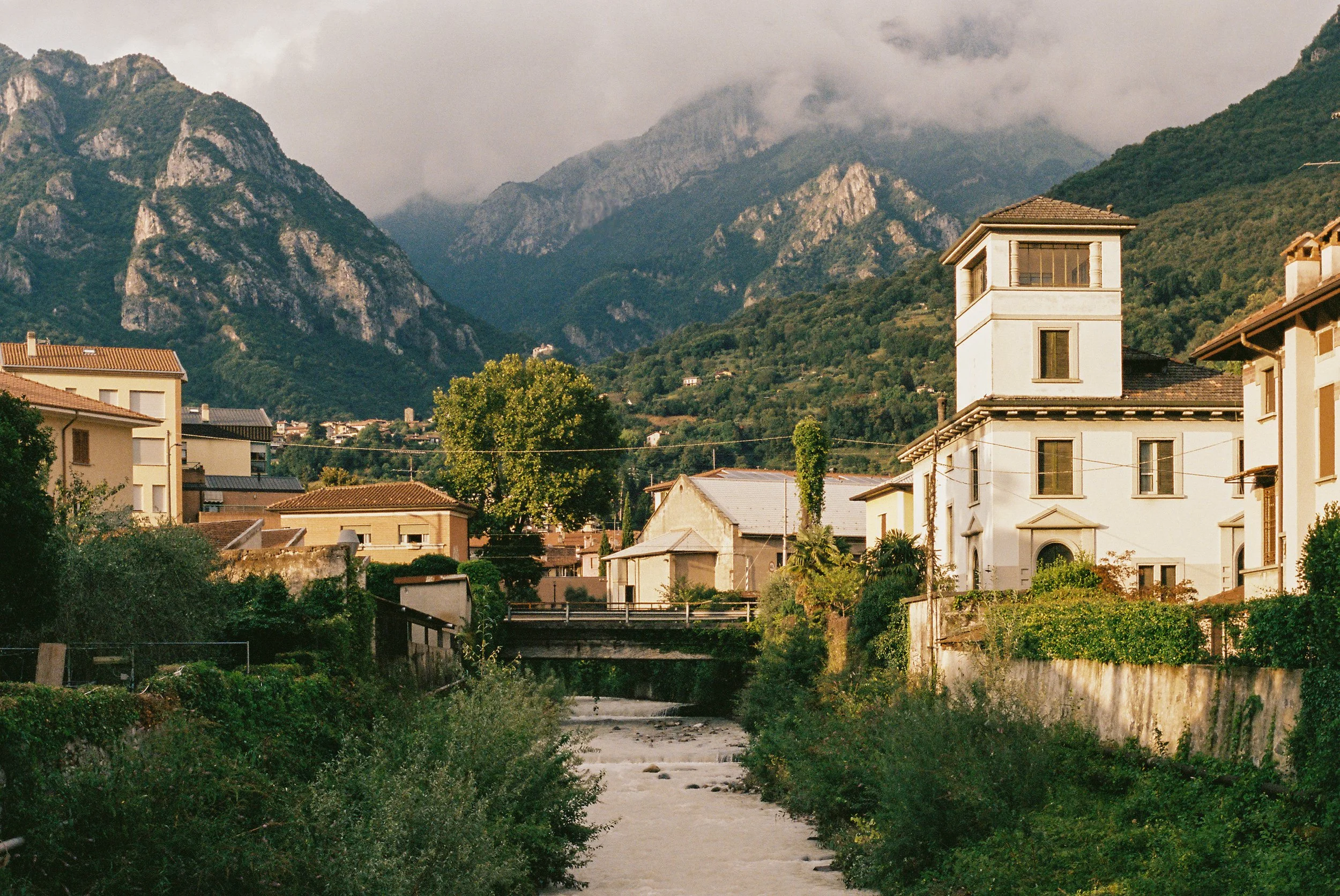 Film photograph of Mandello del Lario village along the river with historic Italian buildings and cloud-covered Alps in the background, Lake Como, Italy — Italy travel photographer and film photographer Anna Hudson