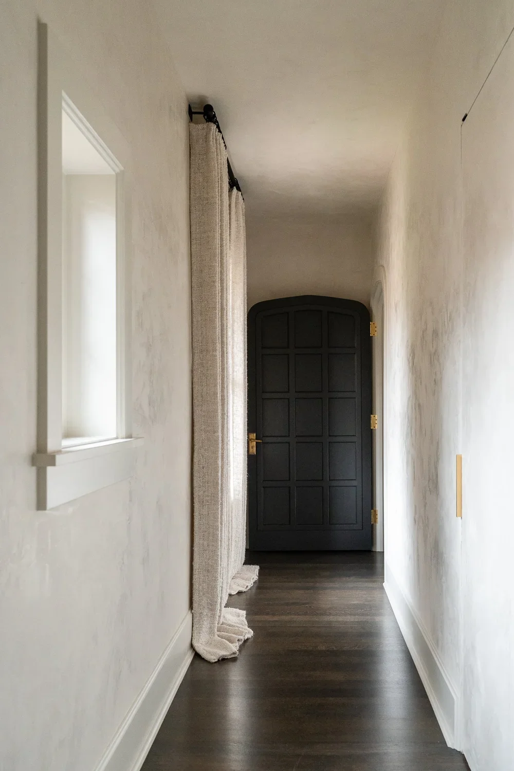 Narrow hallway in historic Tudor home in Cherry Creek, Denver, Colorado with dark hardwood floors, textured plaster walls, linen curtains, and a vintage arched wood door with brass hardware — Denver interior design photographers Anna Hudson