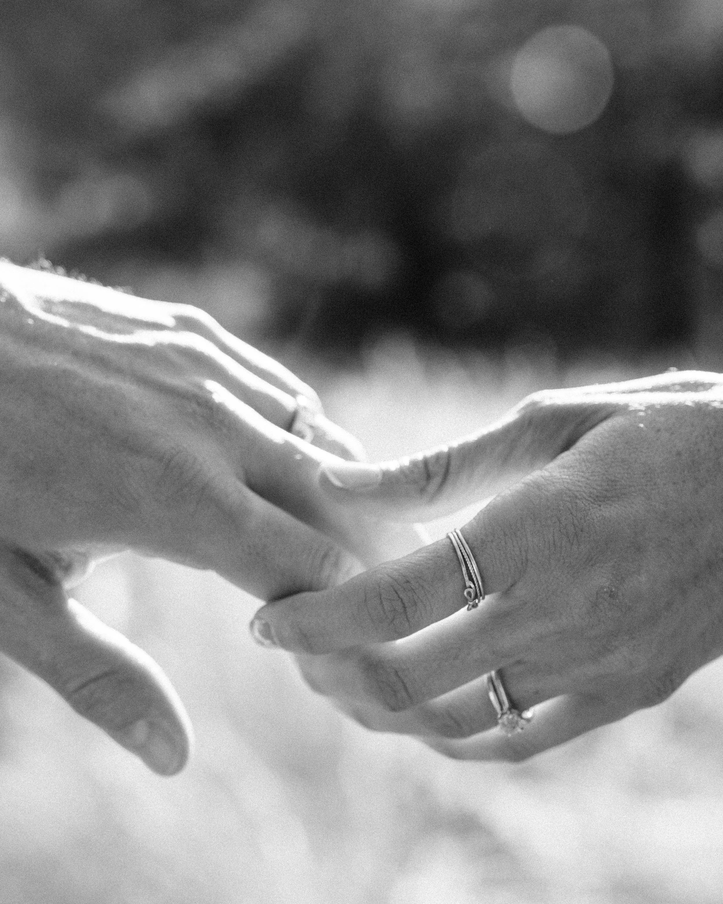 Close-up black and white photograph of a couple holding hands with hand-forged wedding and engagement rings visible, at a Colorado elopement — destination elopement photographer Anna Hudson