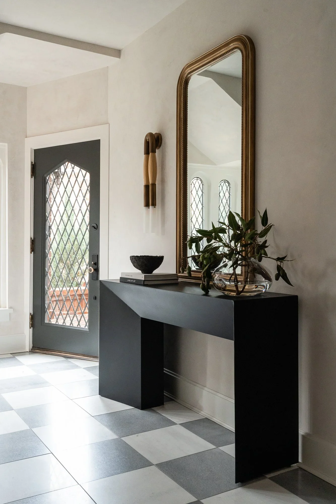 Entryway of a historic Tudor home in Cherry Creek, Denver, Colorado featuring a black console table, gold arched mirror, checkered tile floor, and leaded glass door — Denver interior design photographer Anna Hudson