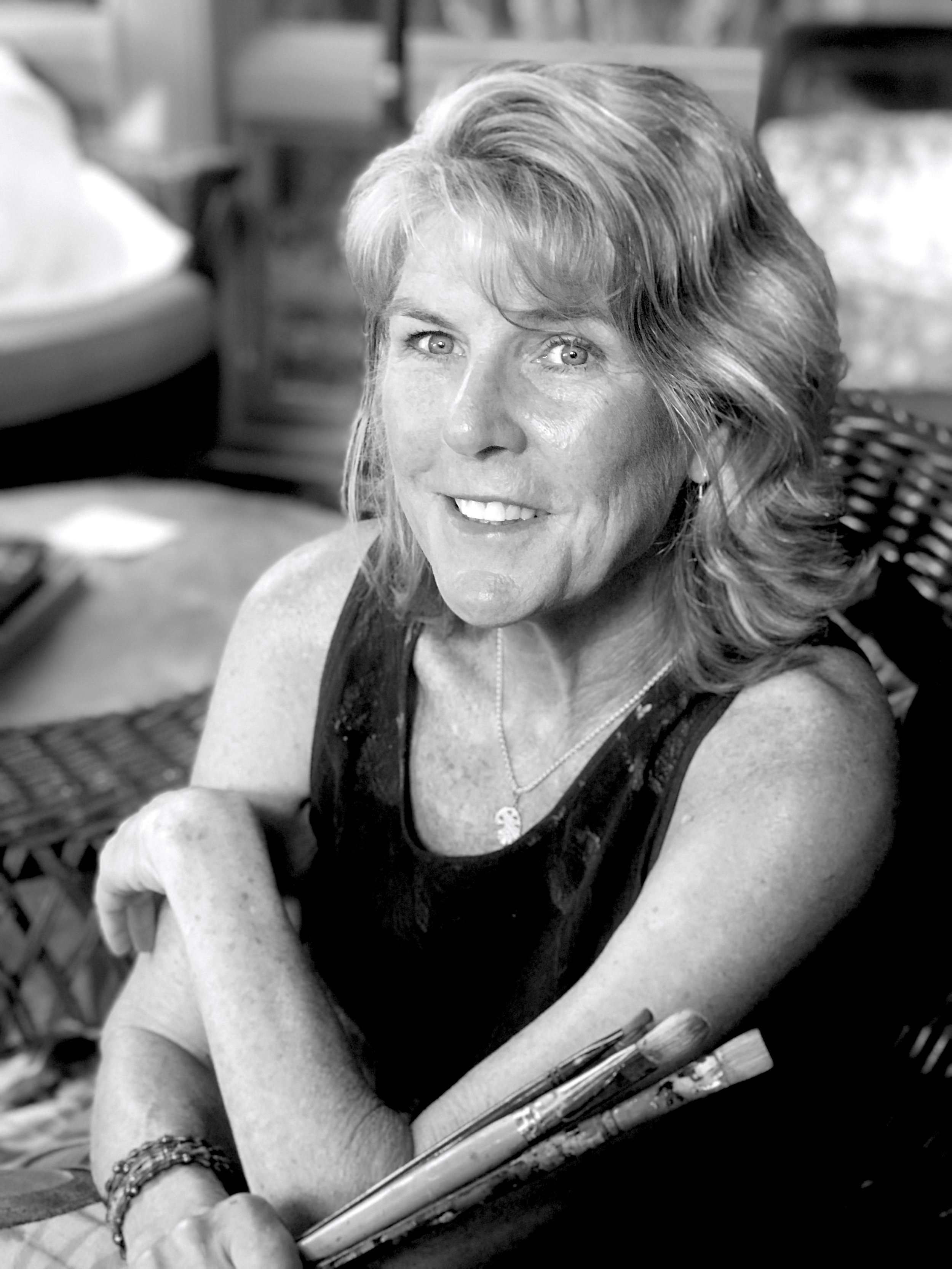 A smiling woman with short, wavy hair sitting outdoors, holding a paintbrush and palette, wearing a sleeveless top and a necklace, in a black-and-white photo.