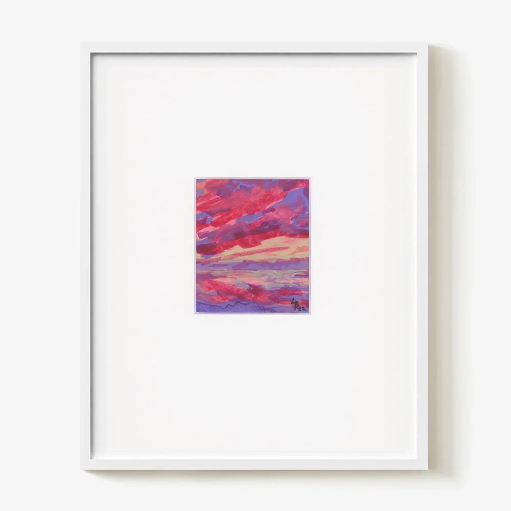 Small colorful painting of a sunset over water framed in white on a white wall.