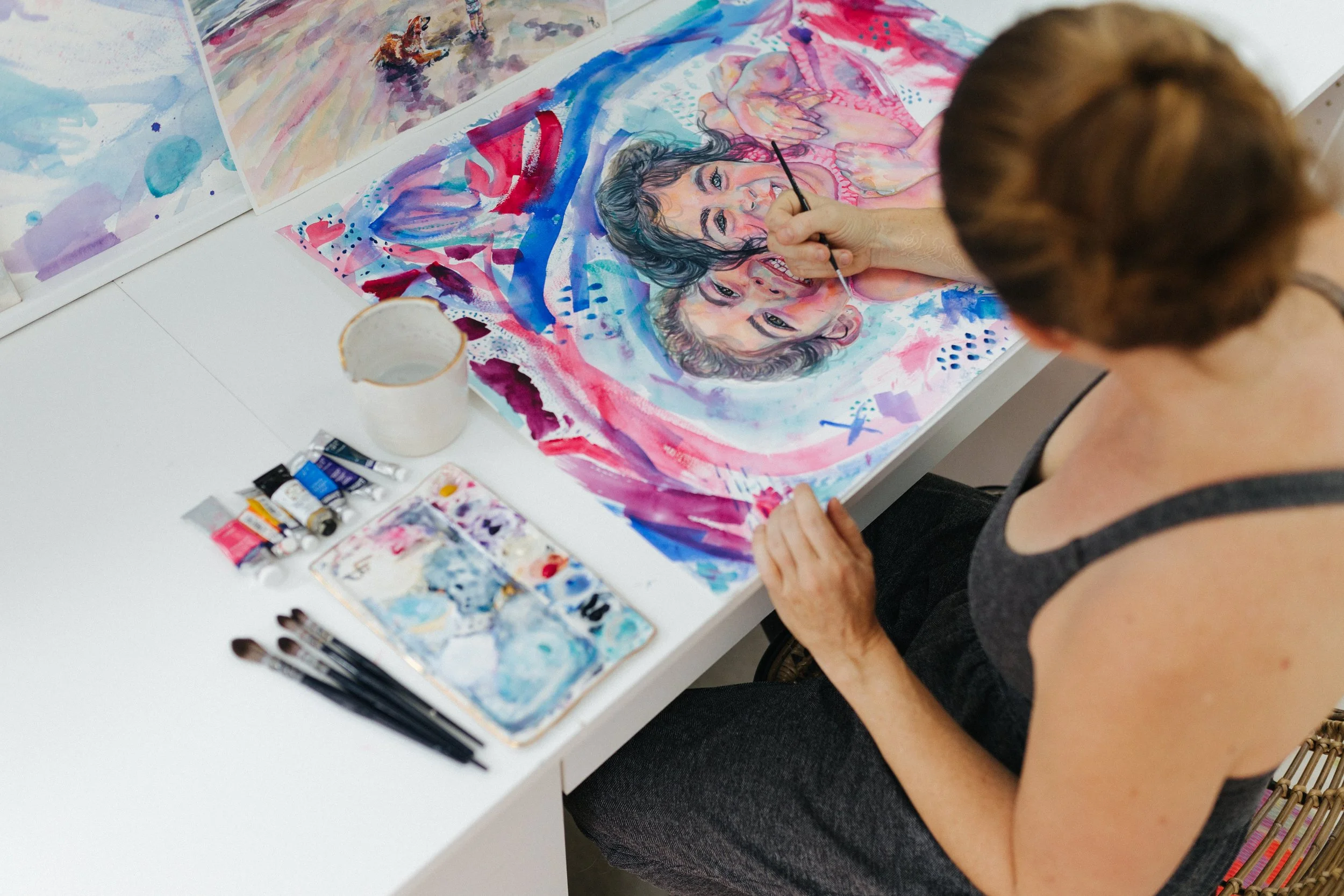 A woman painting a colorful watercolor portrait of two individuals on paper, with various paint supplies and brushes on a white table.