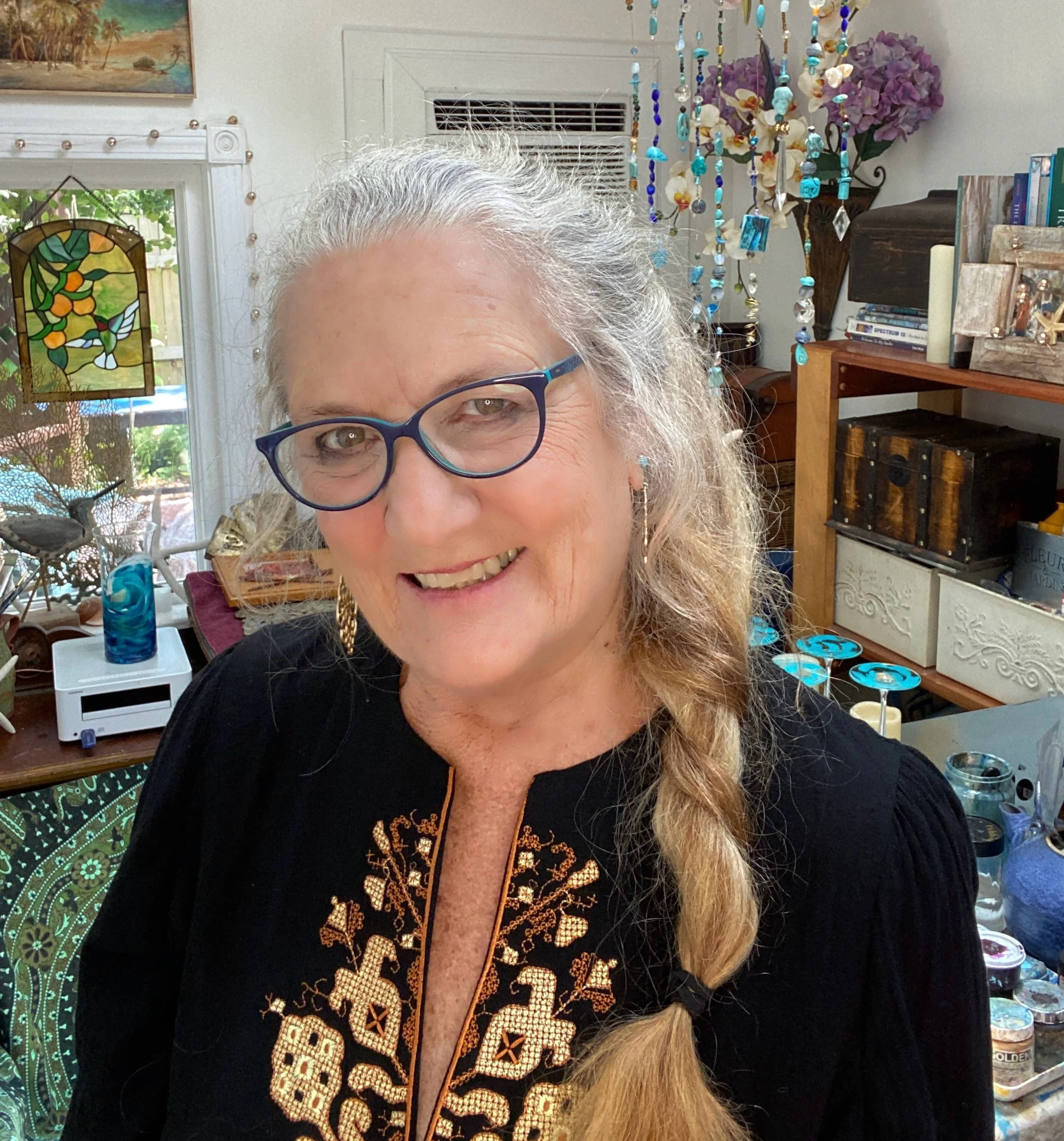 Close-up of a smiling woman with glasses and long curly hair, wearing a black top with gold embroidery, in a room with books, a wooden chest, a stained glass window, hanging bead art, and purple flowers.
