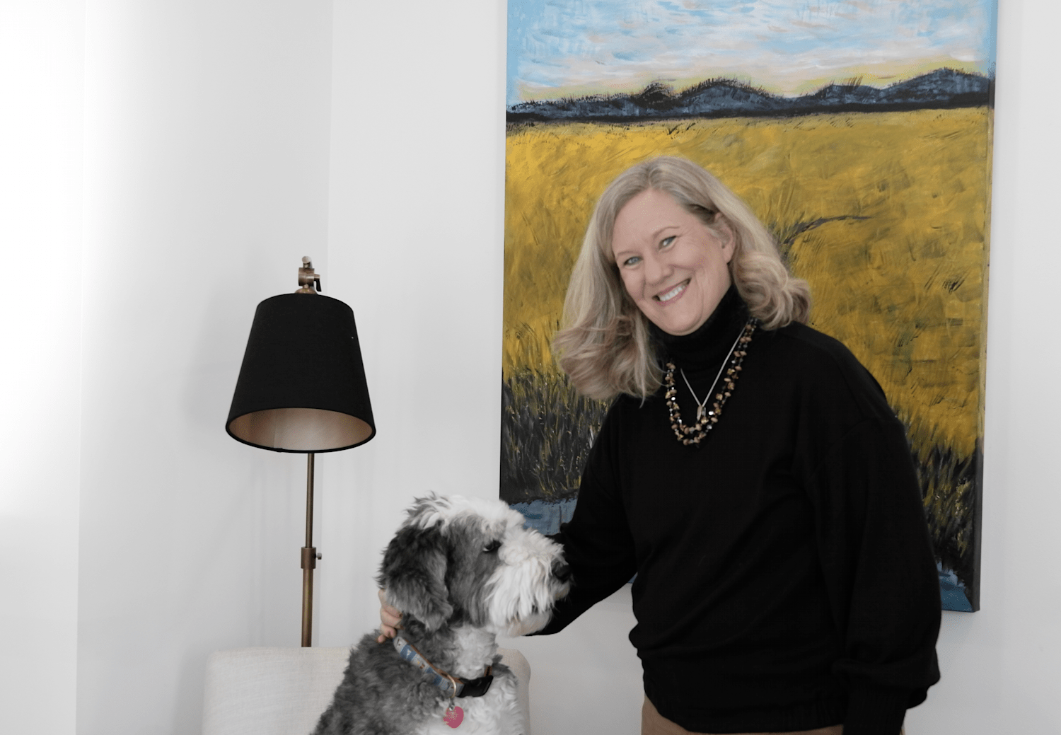 A woman with blonde hair smiling while petting a dog with gray and white fur, in a room with a painting of a landscape and a black lampshade on a floor lamp.