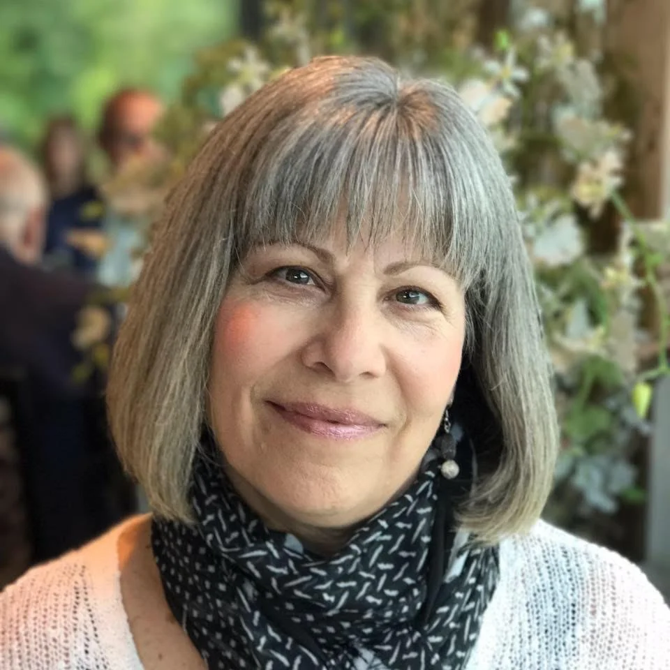 A woman with short gray hair smiling at the camera, wearing a white sweater and black scarf, in a setting with blurred flowers and people in the background.
