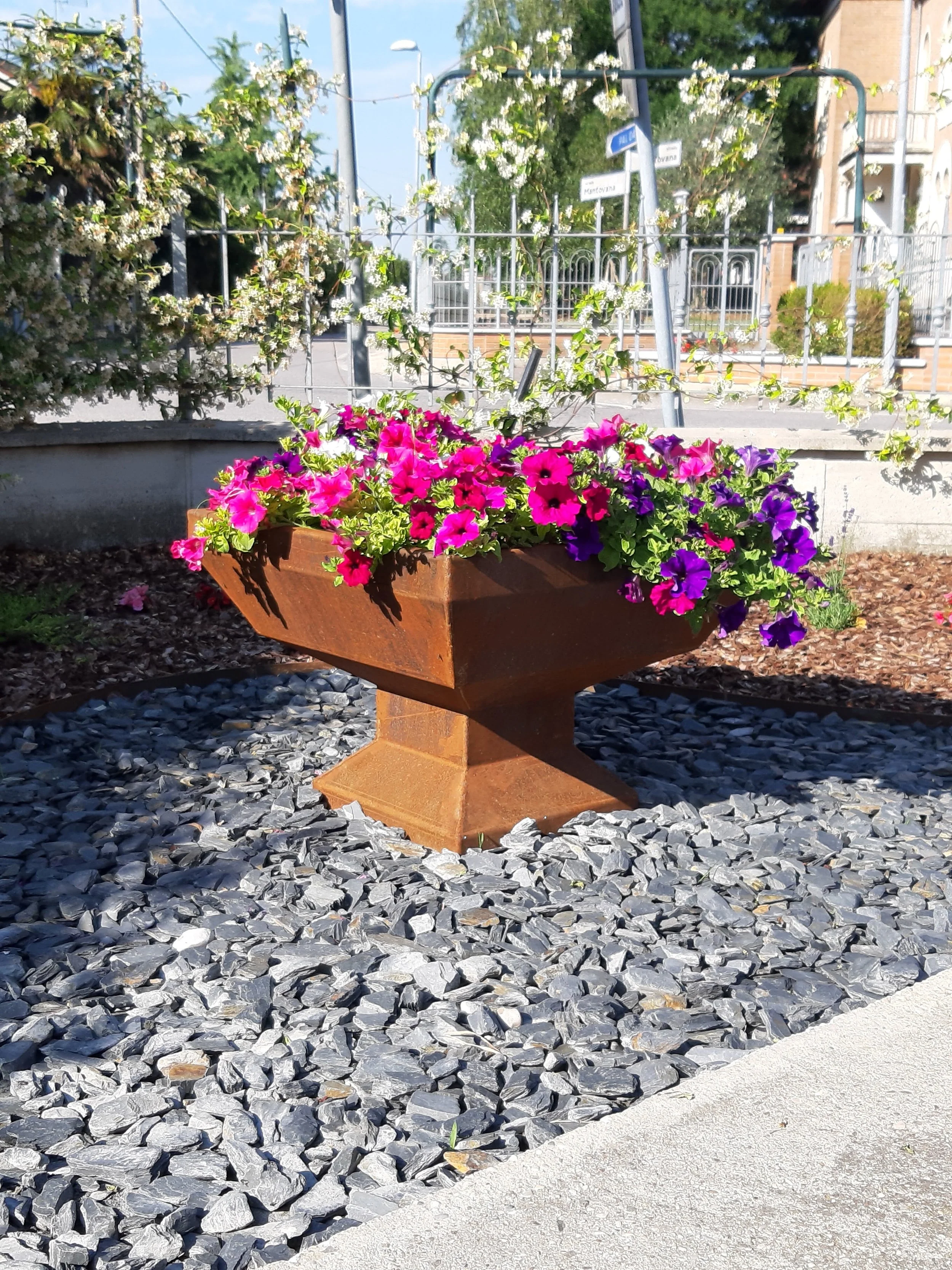 Grande vaso svasato Keystone in acciaio Corten arrugginito, posizionato come elemento centrale in un giardino moderno con fiori colorati.