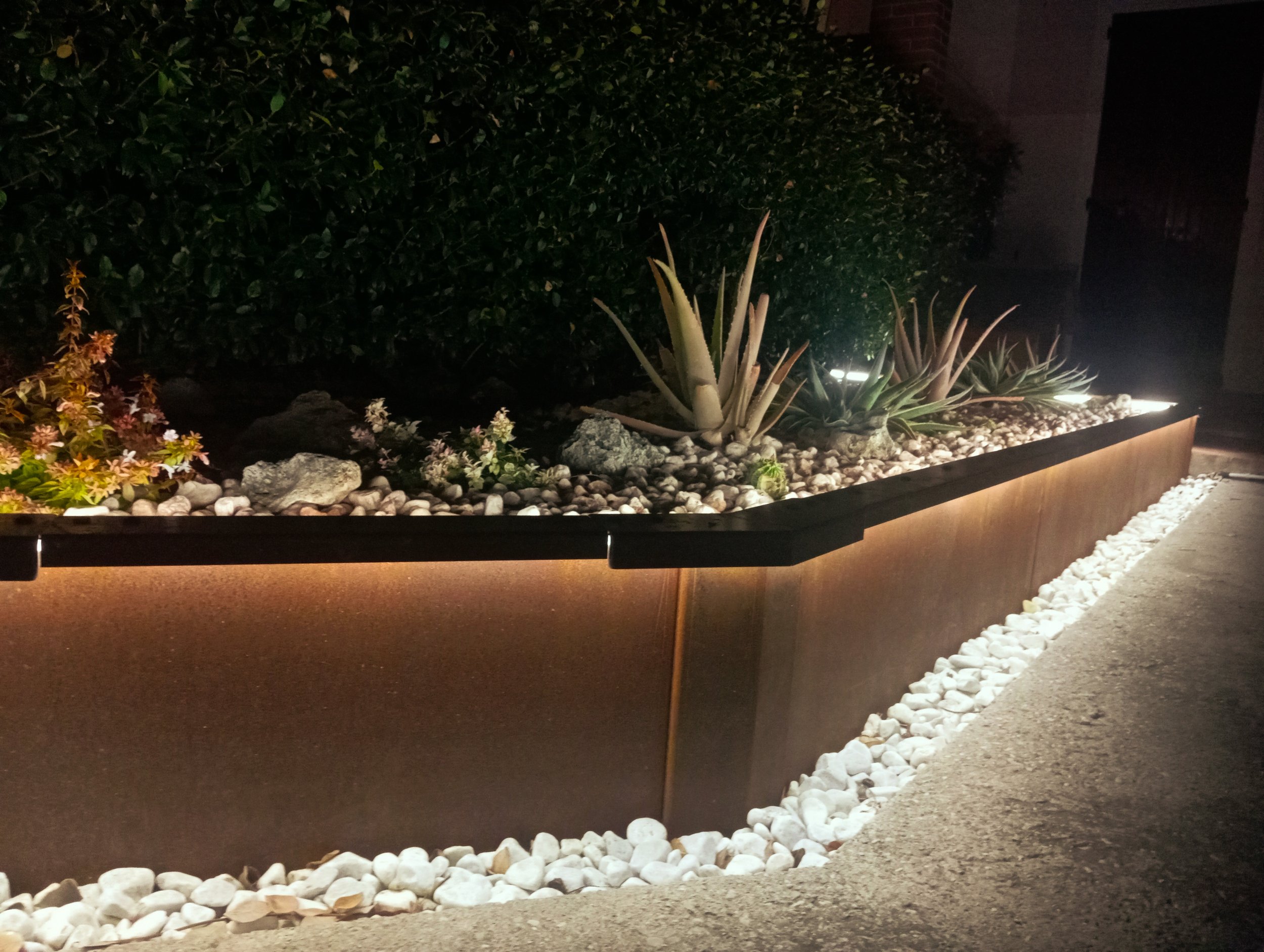 Aiuola rialzata triangolare Vault in acciaio Corten di notte, illuminata alla base da strisce LED che esaltano i sassi bianchi decorativi.