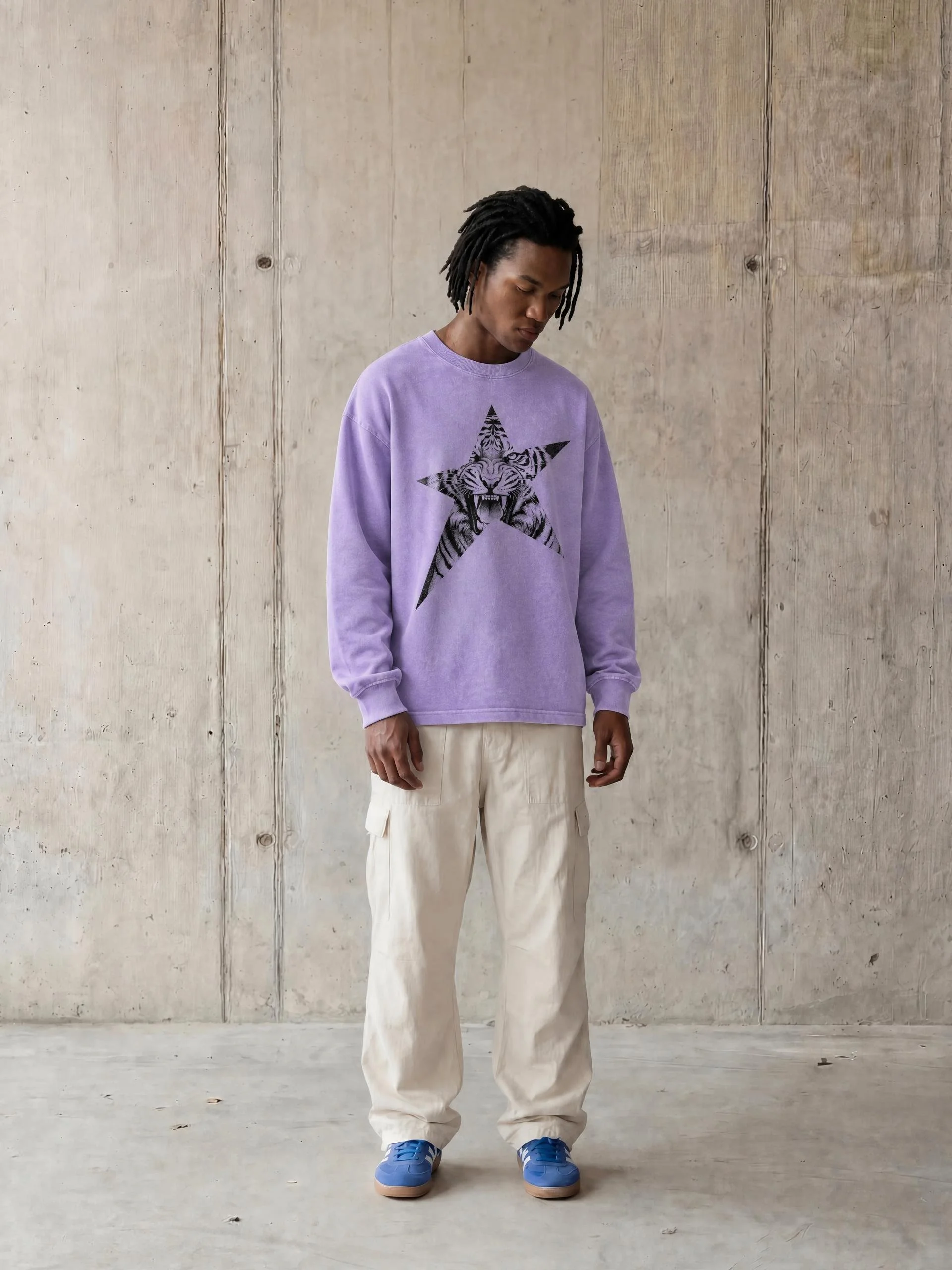 A young man with dreadlocks wearing a purple sweatshirt with a tiger and star design, beige pants, and blue sneakers standing against a plain wooden wall