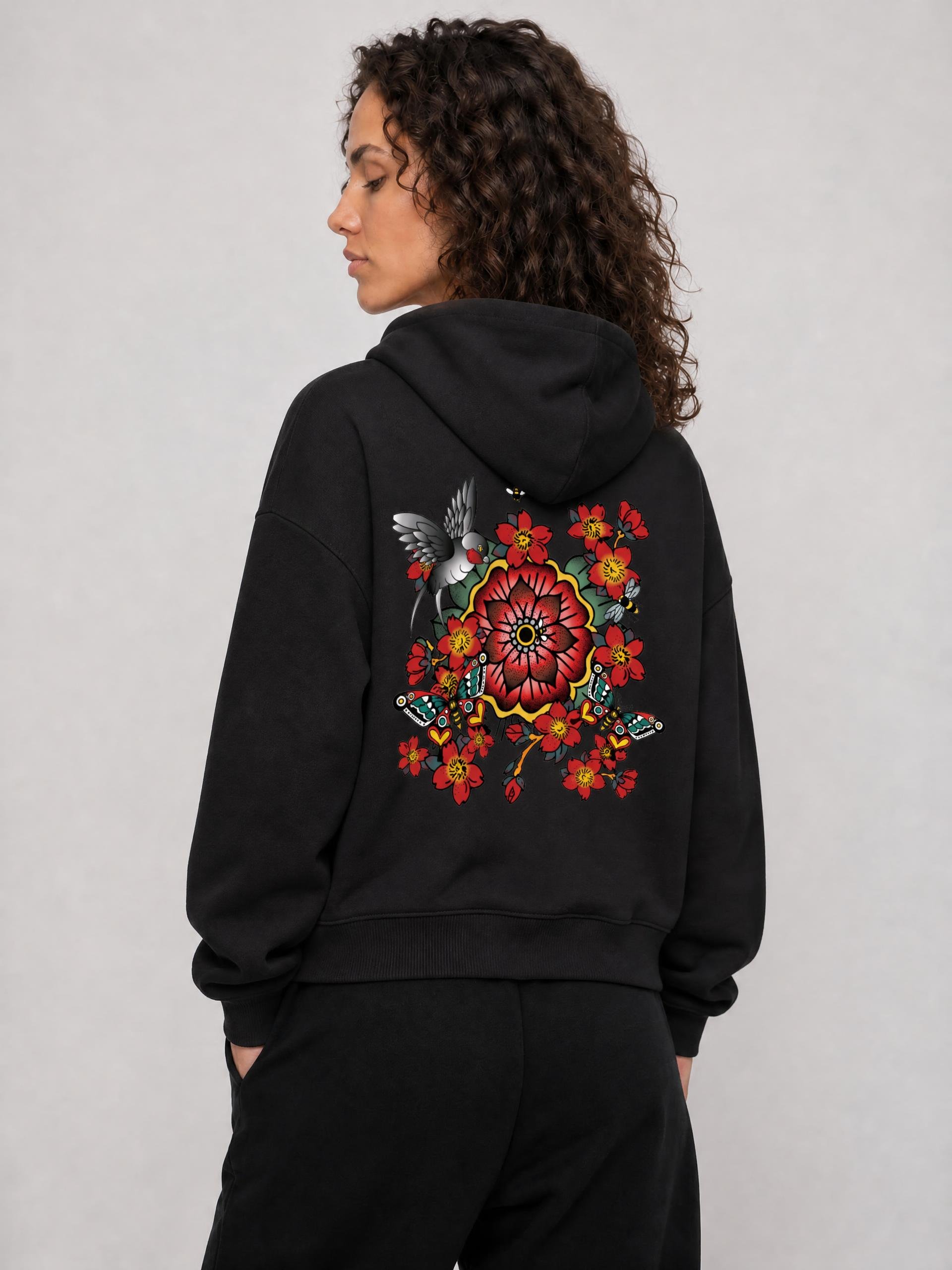 A woman with curly hair wearing a black hoodie with colorful floral and bird embroidery on the back, standing against a plain gray background.
