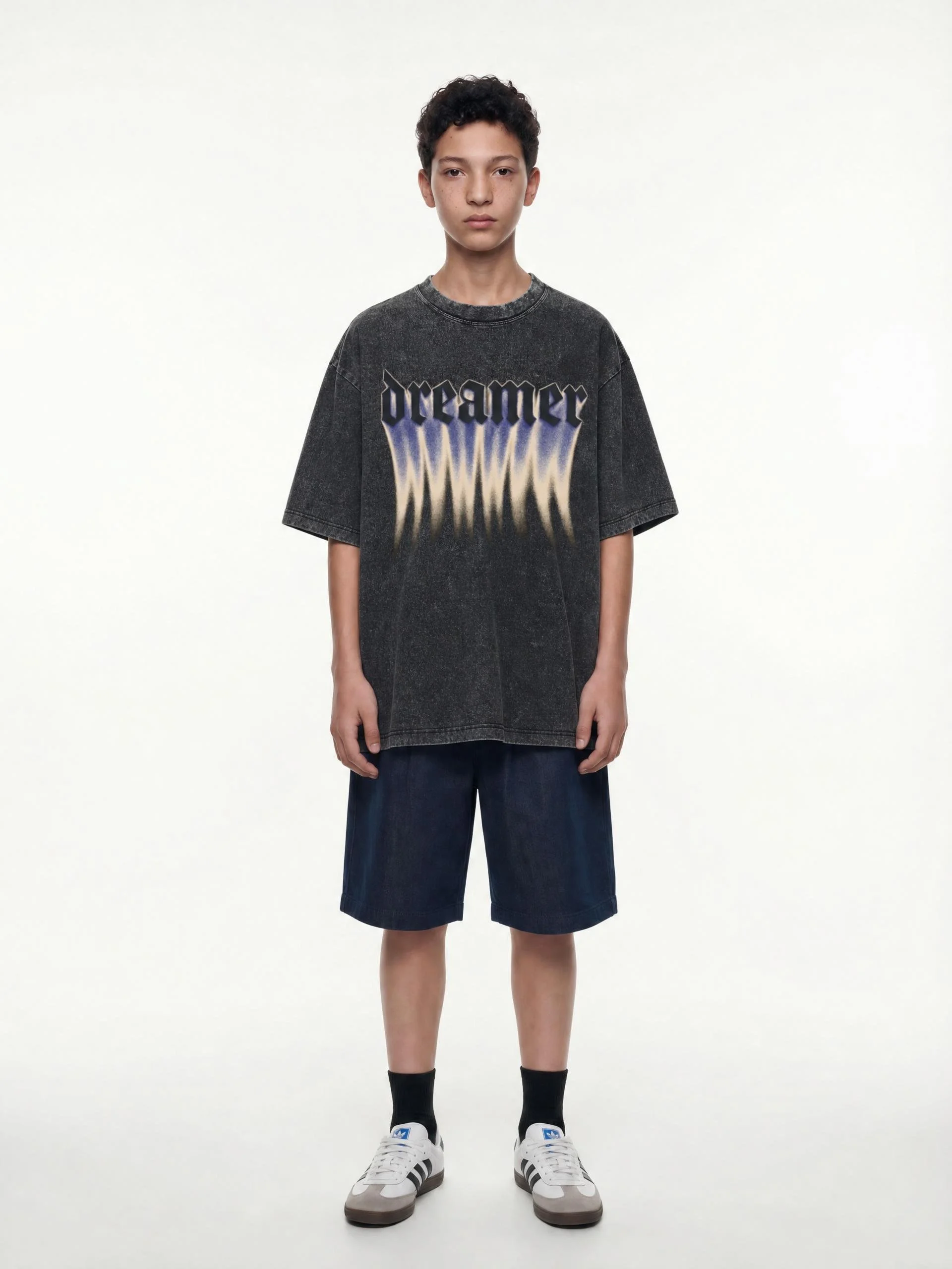 A young person with short dark hair, wearing a black graphic T-shirt that says 'dreamer' in stylized lettering, navy shorts, black socks, and white sneakers, standing against a plain white background.