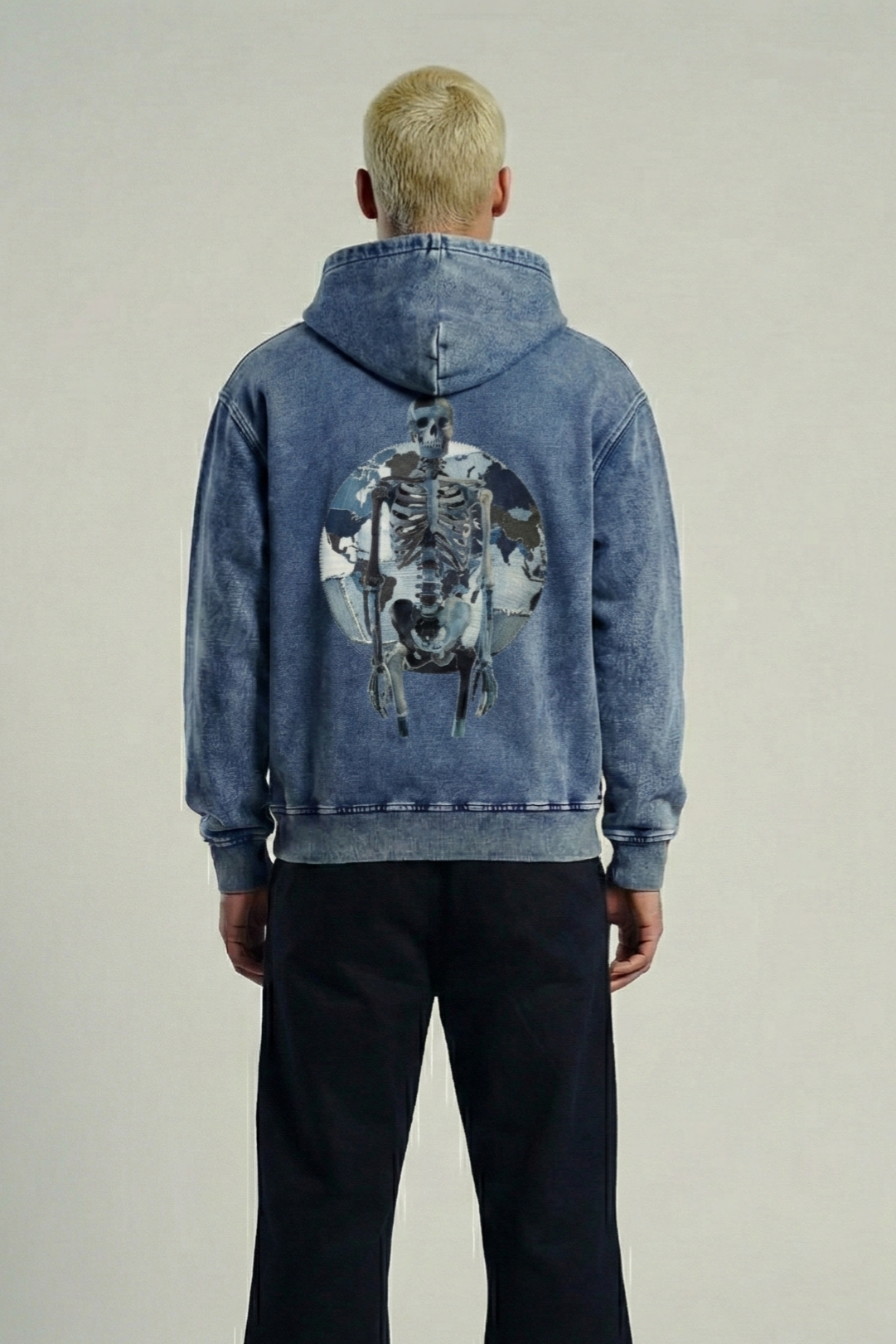 Back of a person with platinum blonde hair wearing a blue denim jacket with a skeleton and dog skull graphic on the back, standing against a plain white wall.