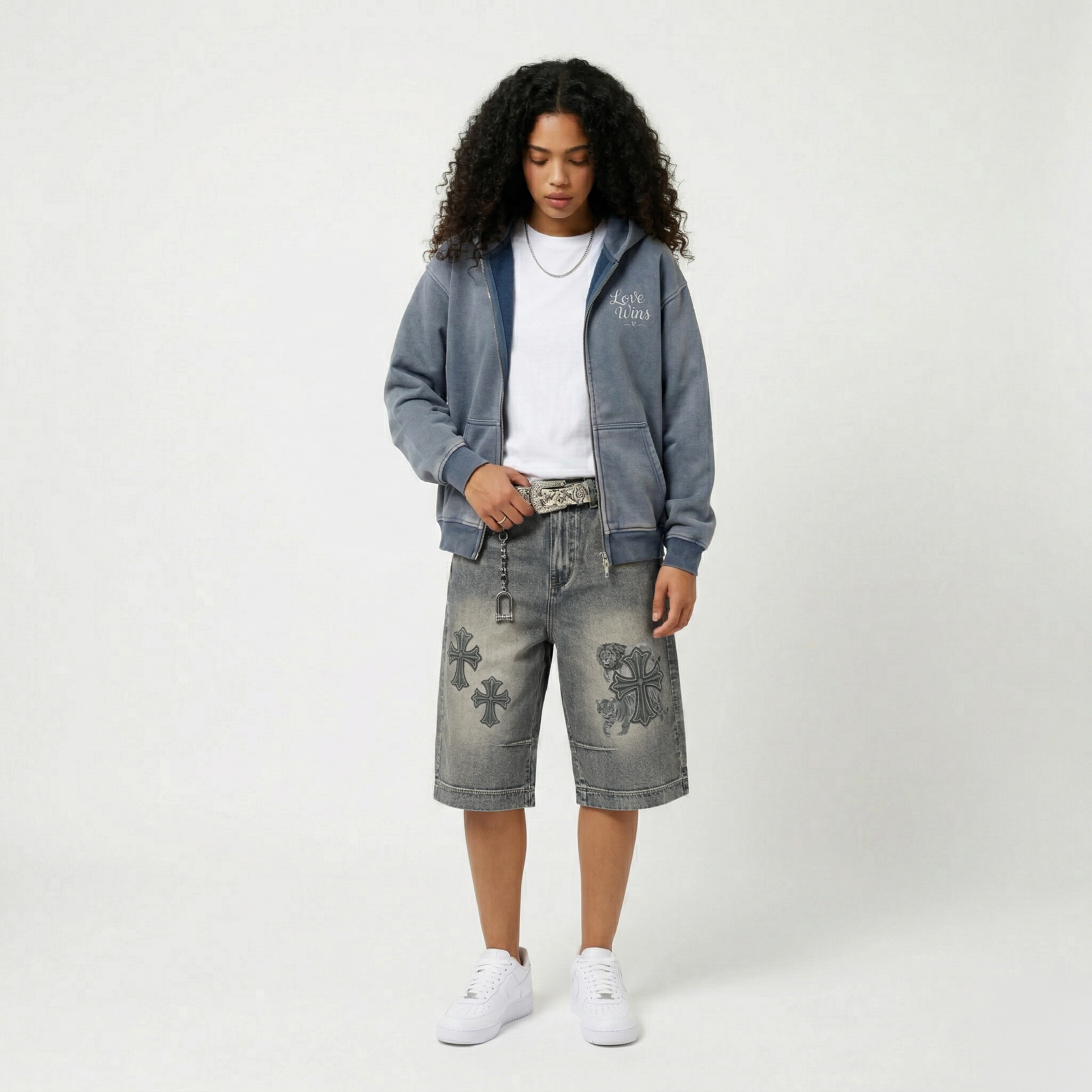A young woman with curly black hair wearing a gray jacket with 'Love Wins' on it, a white T-shirt, patterned denim shorts, white sneakers, and holding a detailed silver belt, standing against a plain white background.