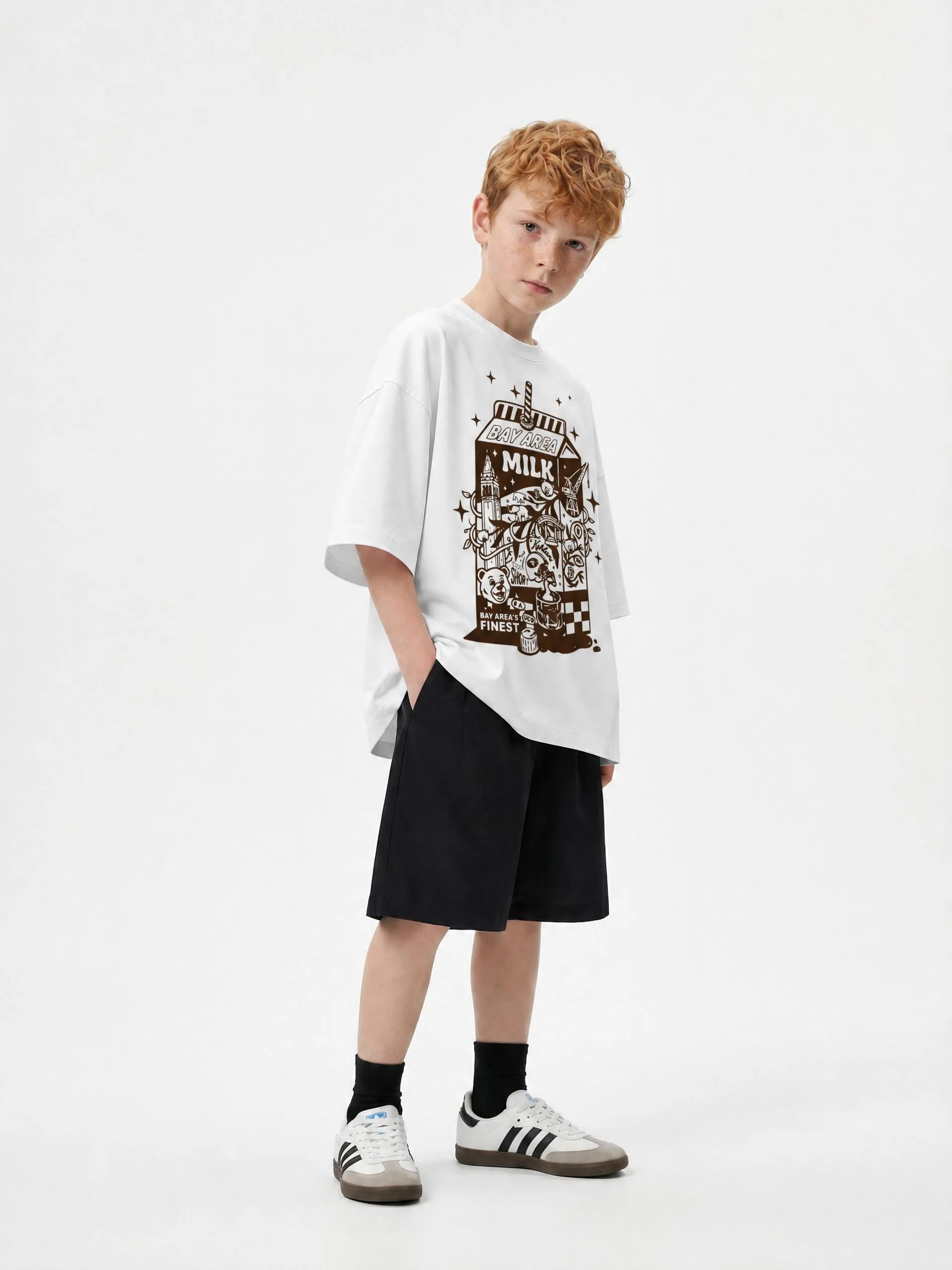 A young boy with curly red hair and light skin, wearing a white oversized graphic T-shirt, black shorts, black socks, and white sneakers with black stripes, posing against a plain white background.