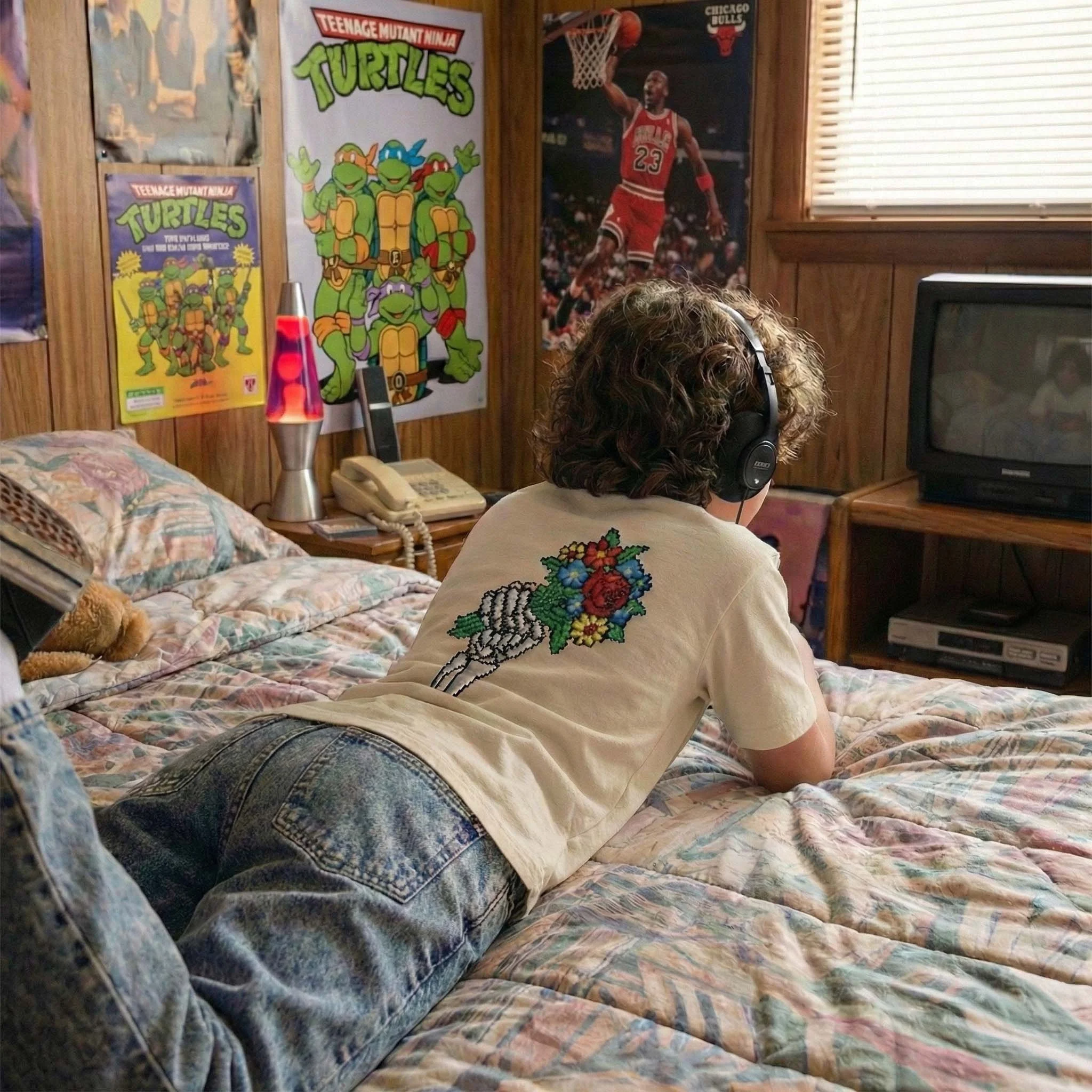 A person with curly hair lying on a bed, wearing headphones and a white T-shirt with a floral embroidery design on the back, watching TV in a room decorated with Teenage Mutant Ninja Turtles posters and a Michael Jordan poster.