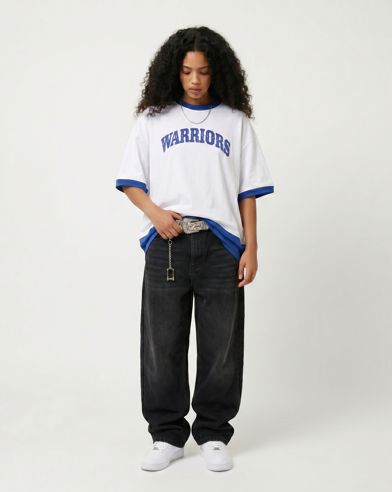 A person with curly dark hair wearing a white and blue hockey jersey with 'WARRIORS' written on it, black wide-leg pants, white sneakers, a silver chain necklace, and holding a silver keychain with a belt buckle, standing against a plain white backgr