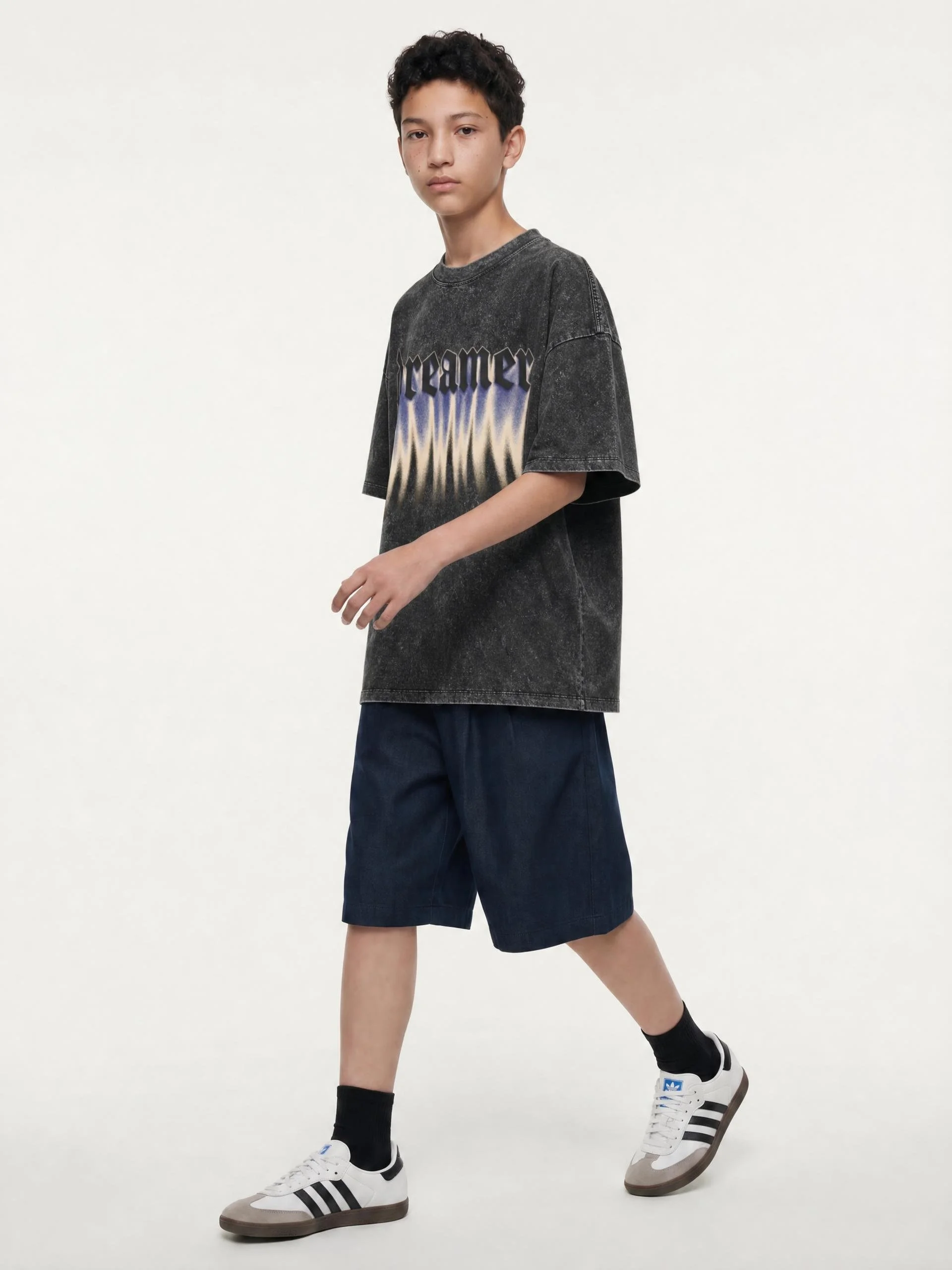 A young boy wearing black shorts, black socks, white Adidas sneakers with black stripes, a black zip-up jacket, and adidas branding, walking against a plain white background.