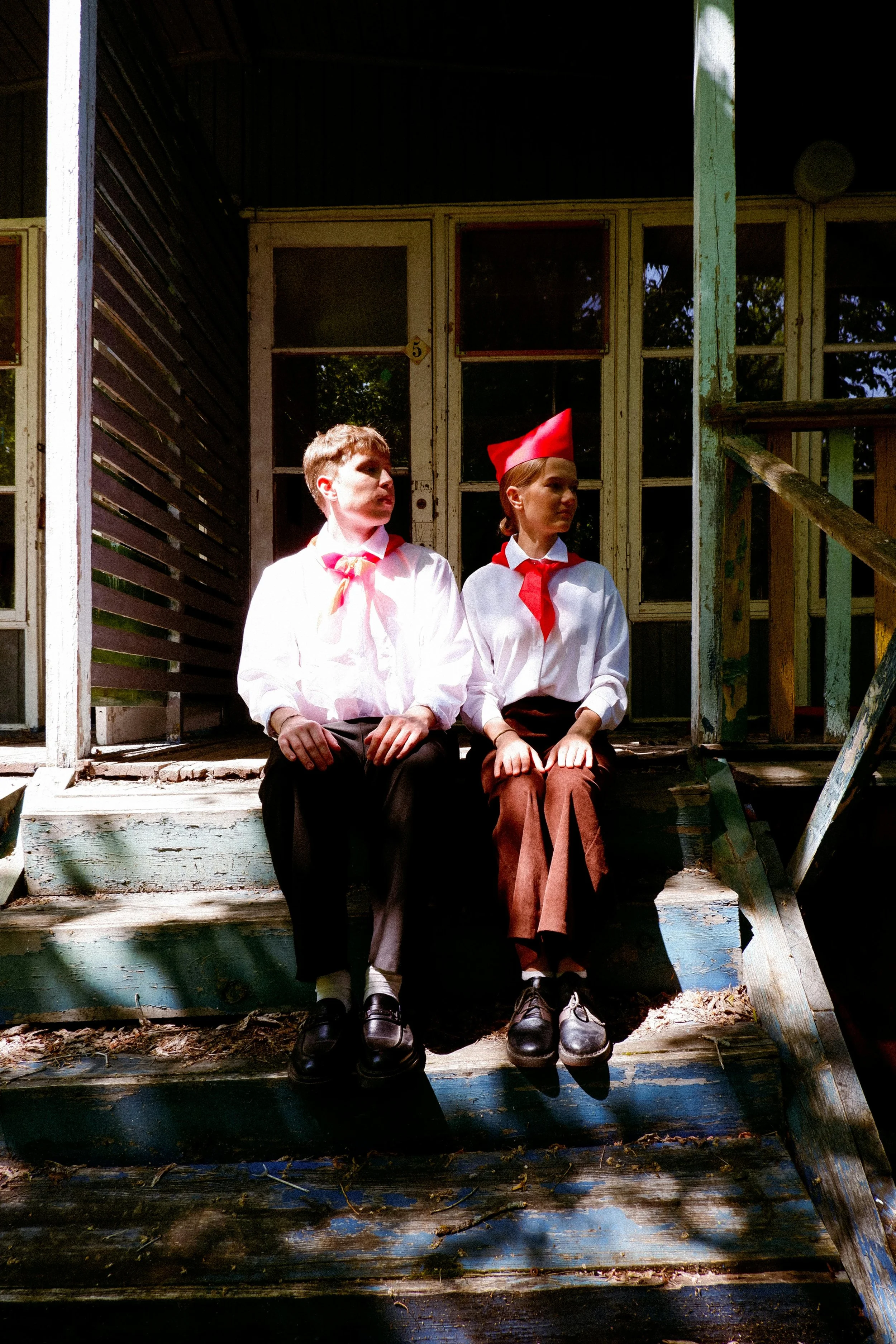 Zwei Jungpioniere in Uniform sitzen auf den Holzstufen eines verfallenen Hauses, dem Sinnbild für Kommunismus und schlechter Führung. Beide schauen in eine Richtung.