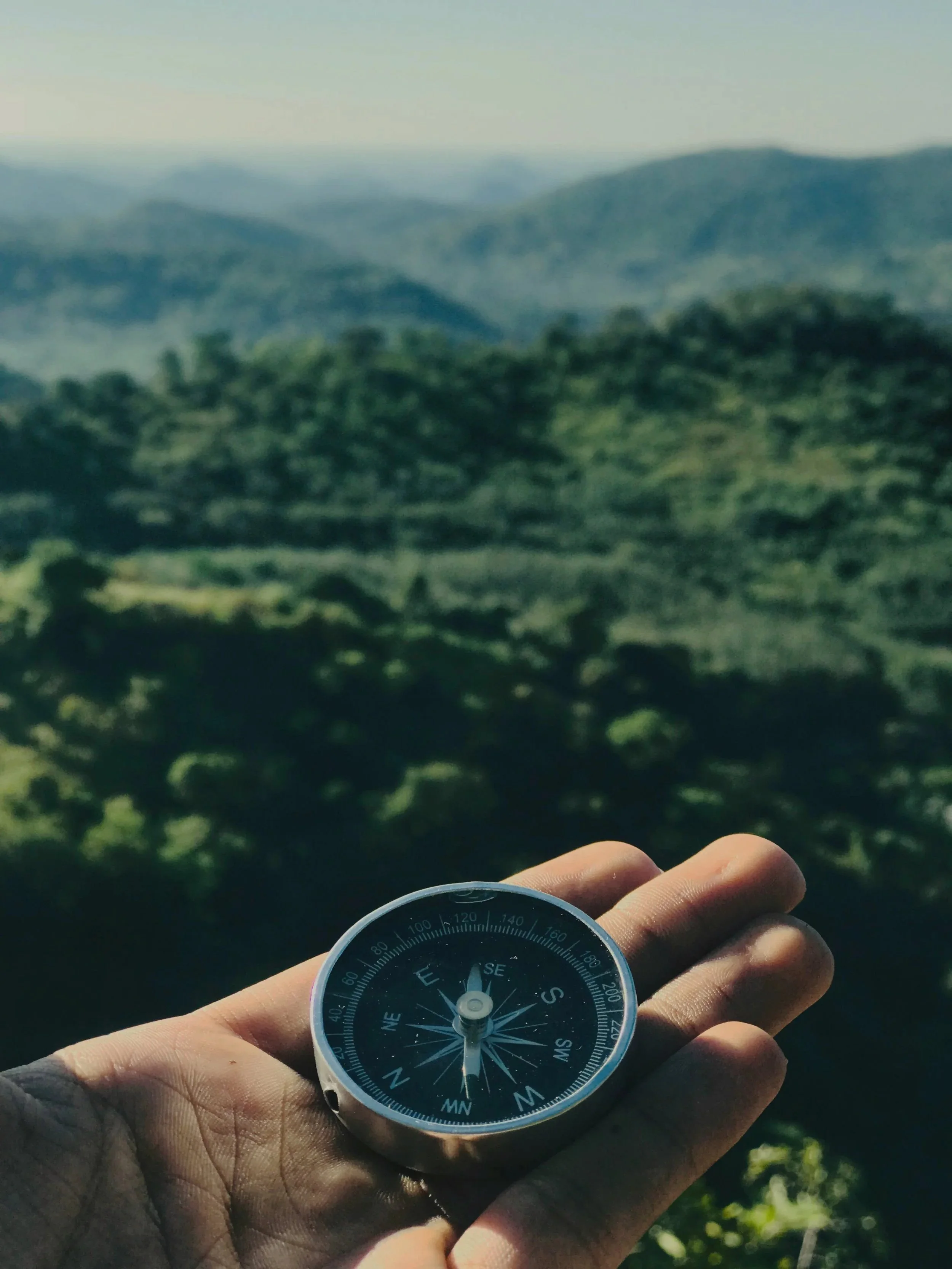 Hand hält Kompass zwecks Orientierung, Überprüfung und systematische Neuausrichtung vor einer grünen, bewaldeten Berglandschaft unter klarem Himmel. Insbesondere für Führungskräfte ist eine systematische Kontrolle des Kurses essentiell.