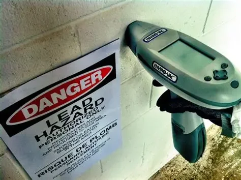 A handheld device used for measuring lead in paint, attached near a wall with a warning sign about lead hazard.