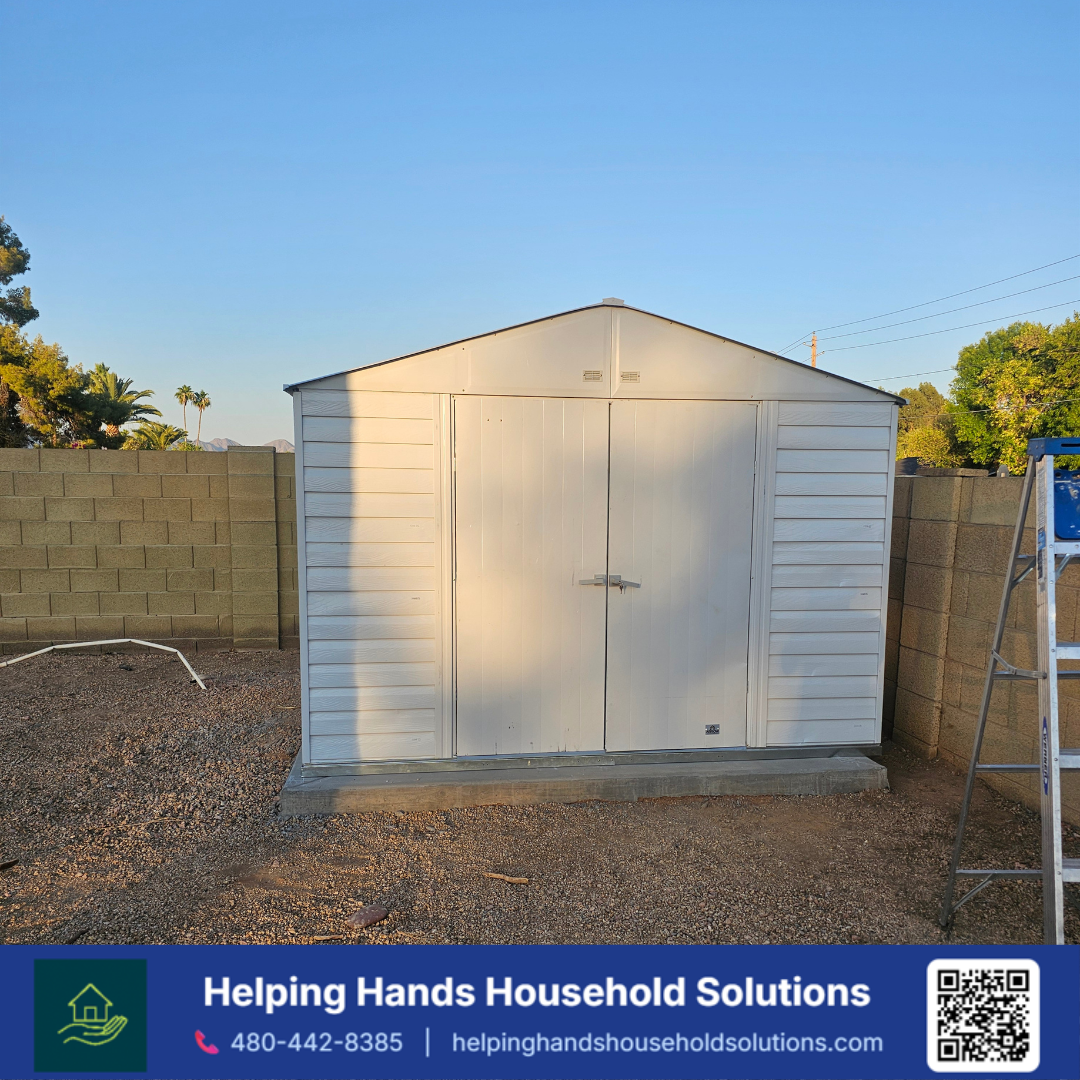Duramax white vinyl storage shed with a peaked shingle-look roof and double doors, installed on a paver base along a backyard block wall in Phoenix, AZ by Helping Hands Household Solutions — clean, minimalist panel design offering low-maintenance, we