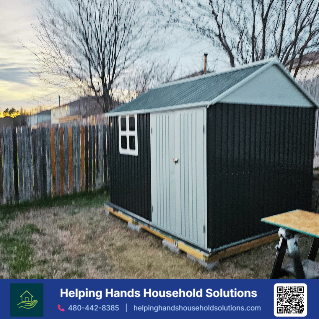Two-tone steel garden shed with light gray gable ends, a matching corrugated roof, and dark green corrugated side panels, installed on a wood skid base in a Phoenix, AZ backyard by Helping Hands Household Solutions — classic gable-roof design deliver
