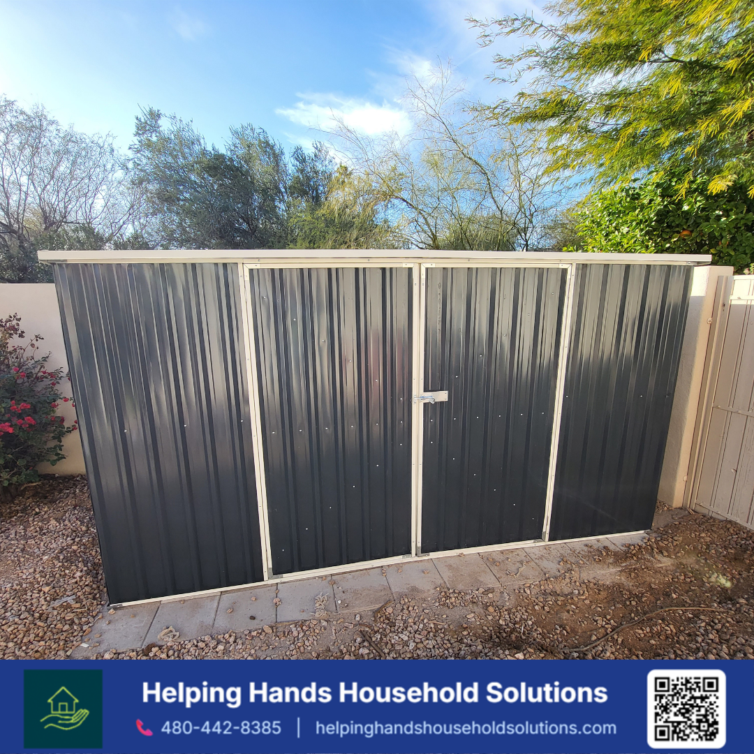 Absco charcoal corrugated steel storage shed with a slanted lean-to roof and cream trim accents, installed on a gravel base along a backyard privacy wall in Phoenix, AZ by Helping Hands Household Solutions — rugged Australian-engineered steel constru