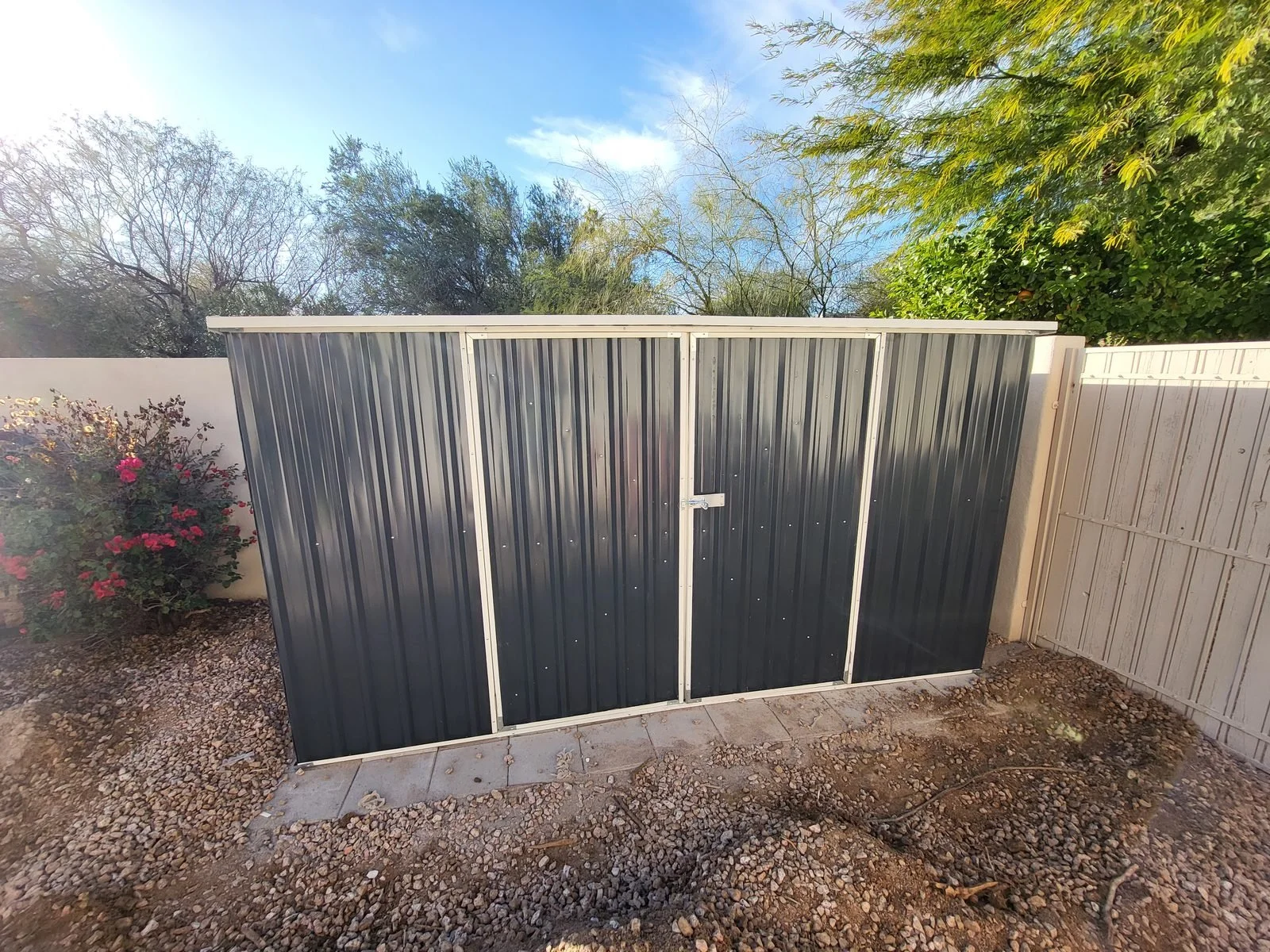 Long single-slope metal storage shed with double doors assembled along a backyard wall in the Phoenix, AZ area by Helping Hands Household Solutions