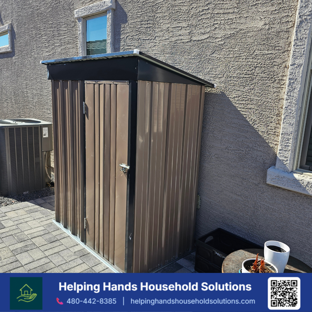 Narrow tan corrugated steel storage shed with a black slanted roof and matching door frame, installed against a stucco exterior wall in a Phoenix, AZ backyard by Helping Hands Household Solutions — compact vertical design maximizes storage in tight s