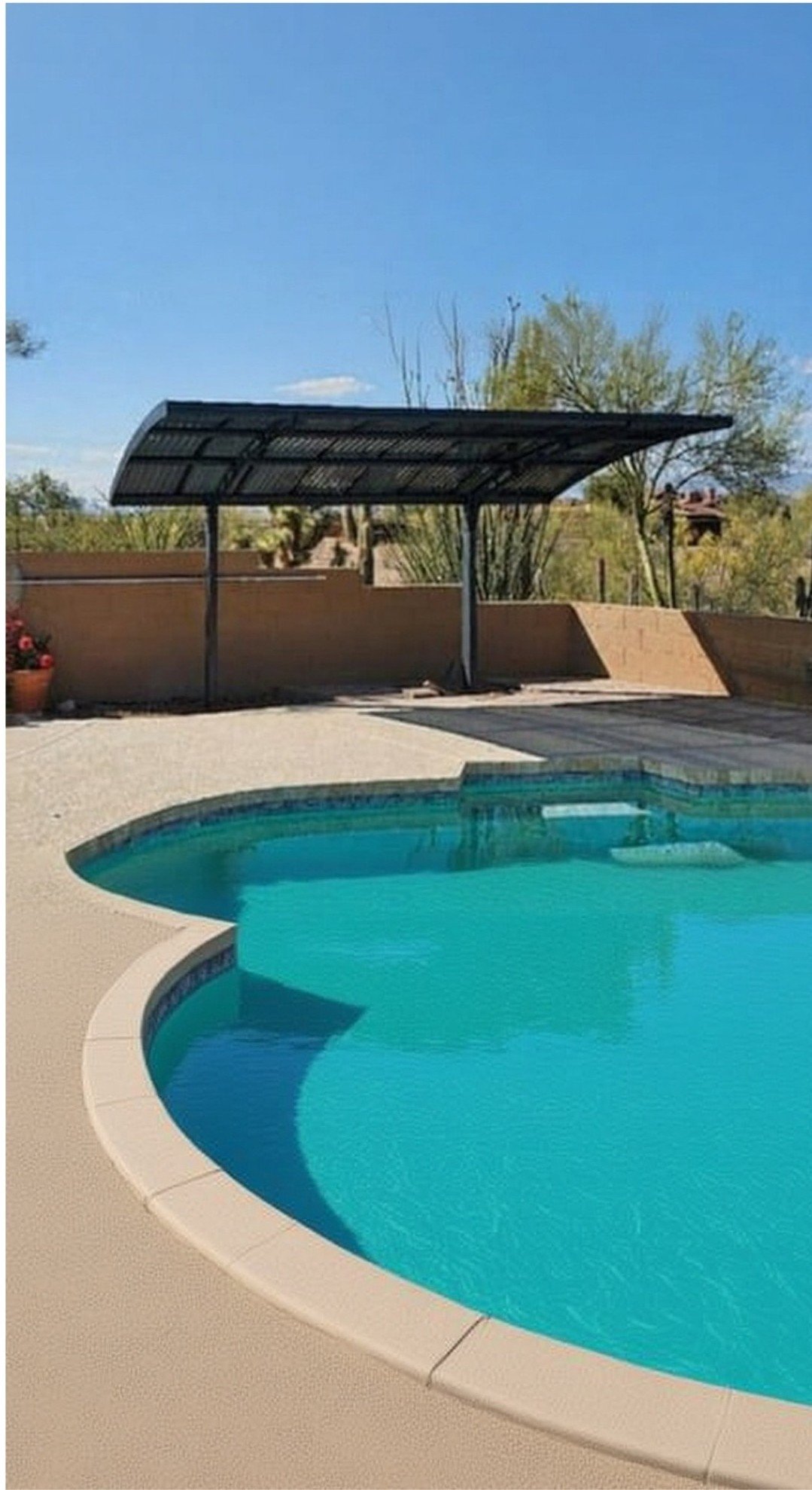 A swimming pool with a rounded edge, surrounded by a concrete deck, under a clear blue sky with a carport shade structure and desert plants in the background.