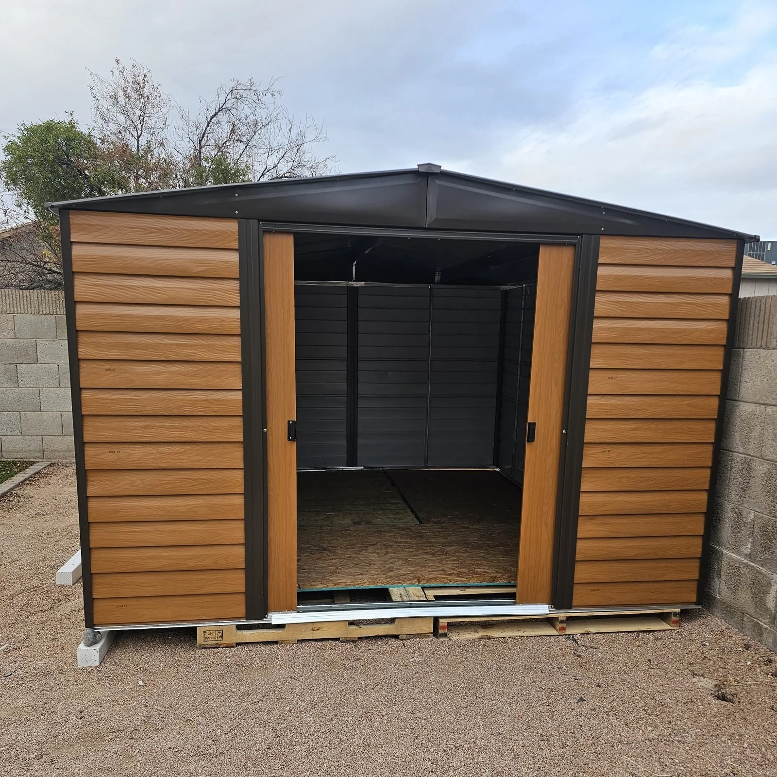 Wood-grain metal storage shed with sliding doors assembled in a Phoenix, AZ backyard by Helping Hands Household Solutions