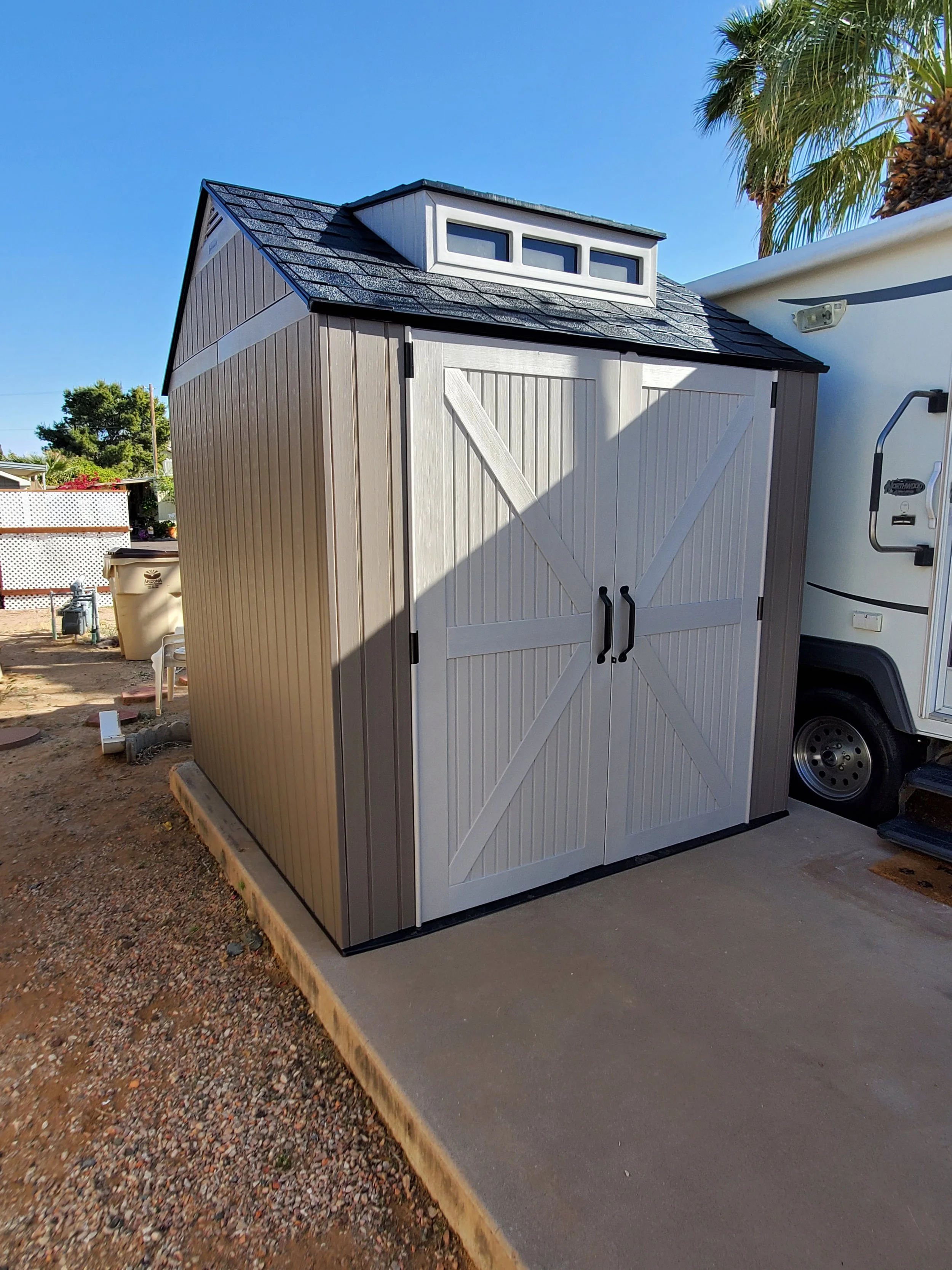 Lifetime two-tone resin storage shed from Costco with diagonal-plank double doors, round handles, and a dormer roof vent, installed on a concrete pad in a Phoenix, AZ backyard by Helping Hands Household Solutions — durable, low-maintenance constructi