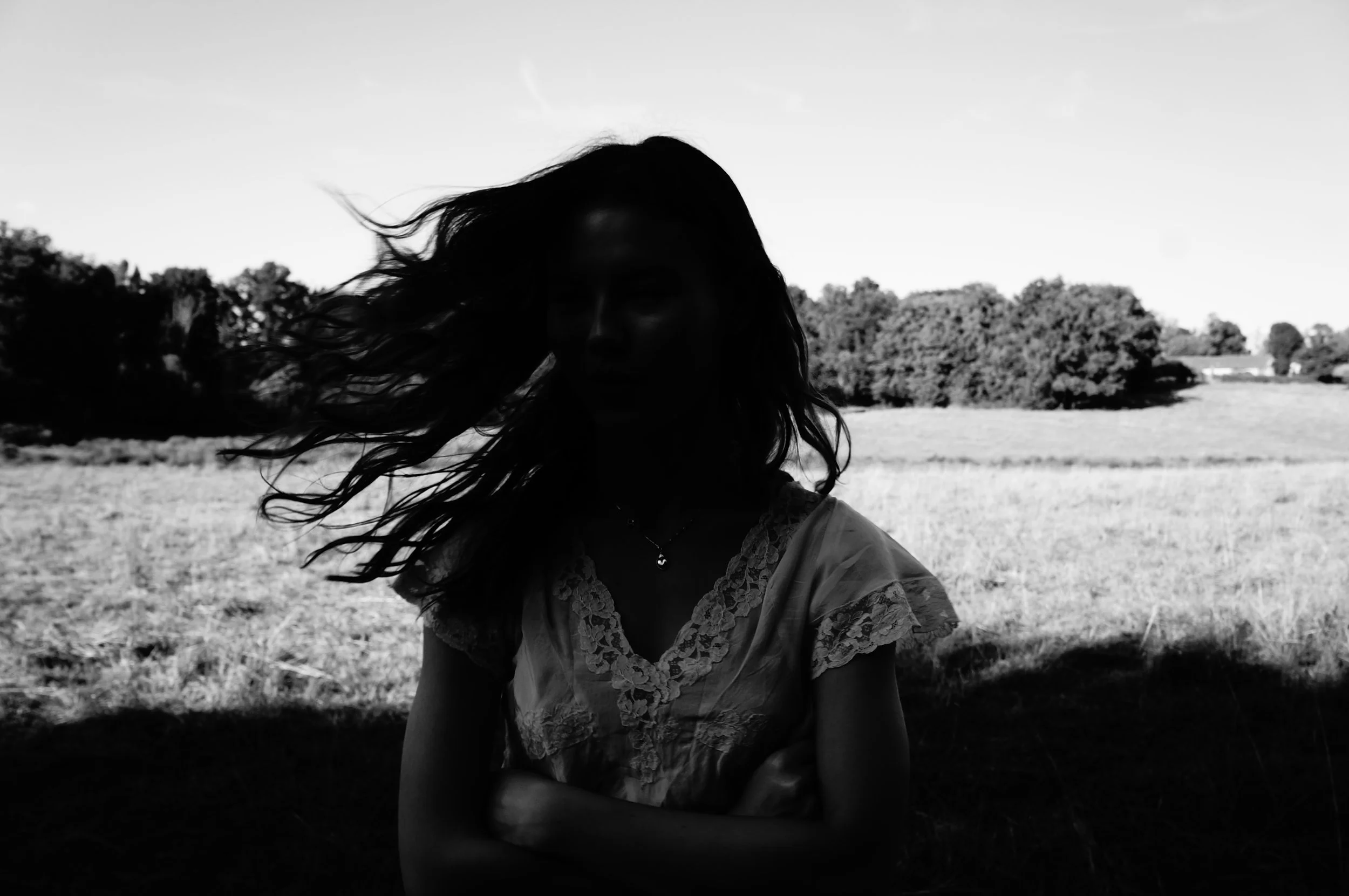 A woman with long hair standing outdoors in a field, shadows on her face, with trees in the background.