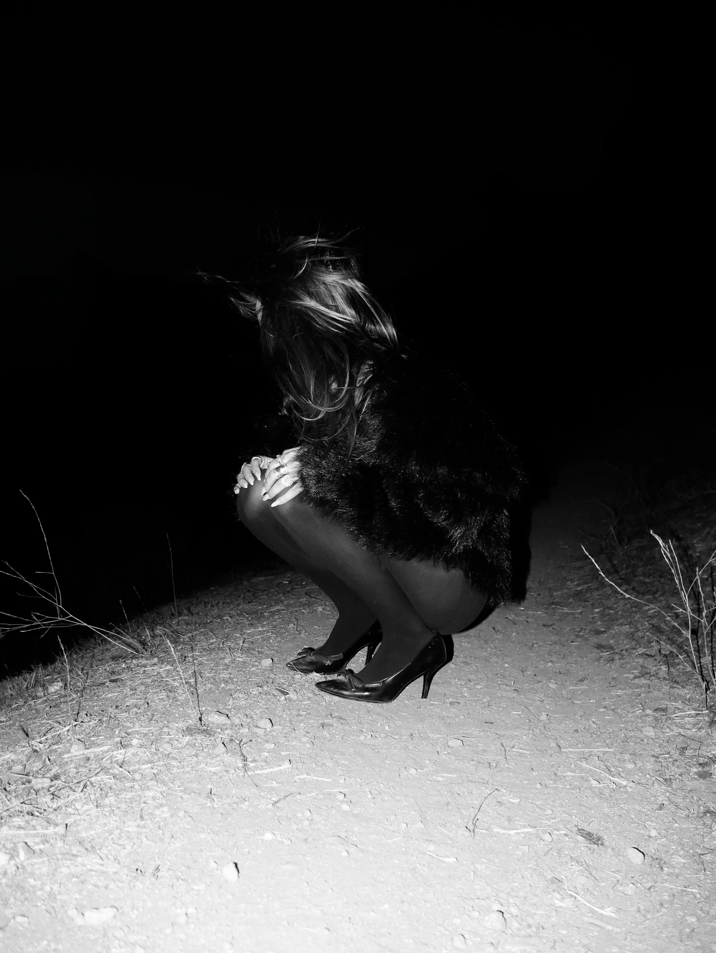 A woman squatting outdoors at night, dressed in high heels, tights, a fur coat, and with long hair, illuminated by a bright light, against a dark background.