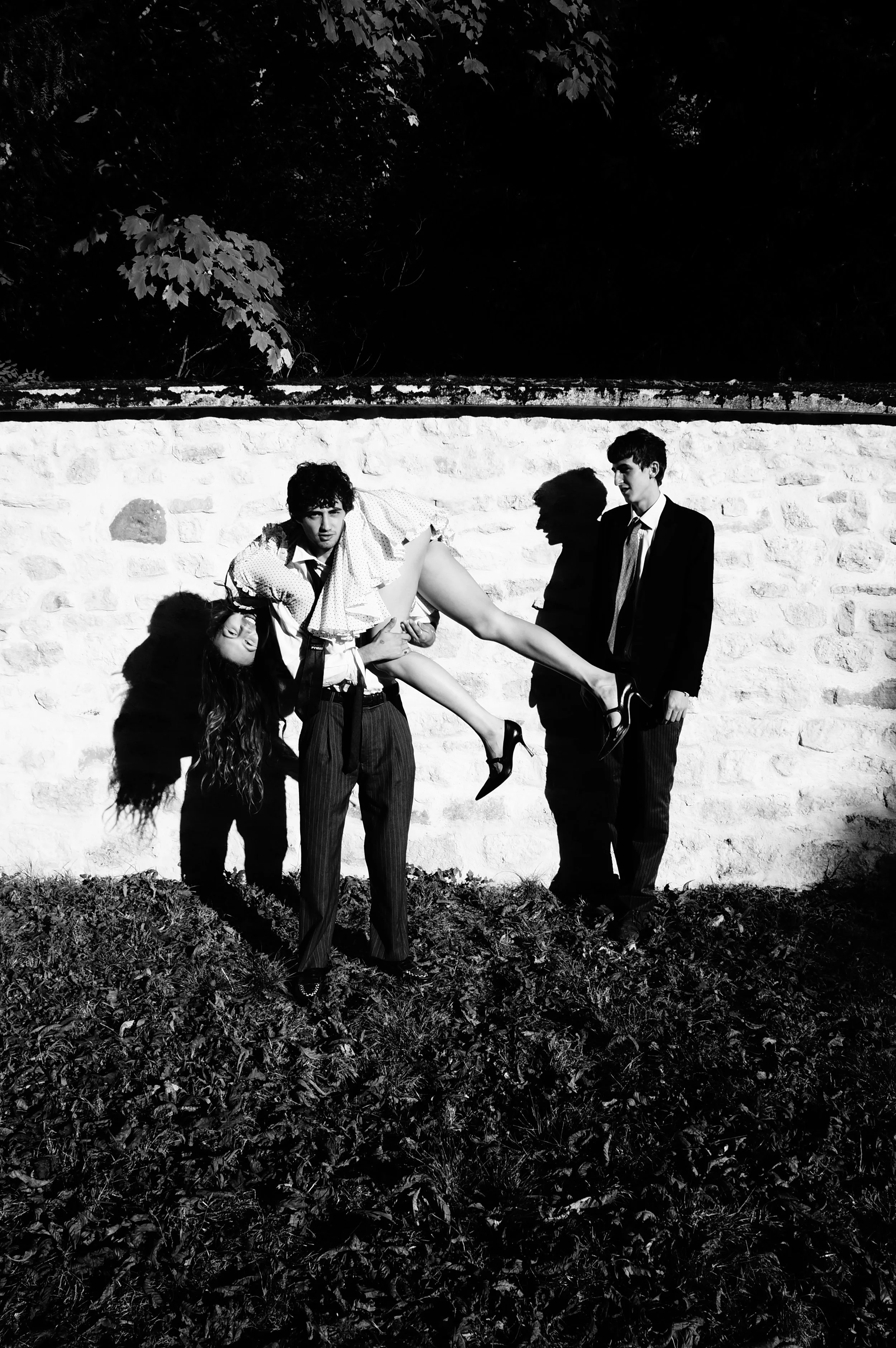 Three people, two men and a woman, posing outdoors against a brick wall. One man is holding the woman upside down by her waist, and her head is hanging down near the ground. The scene is in black and white.