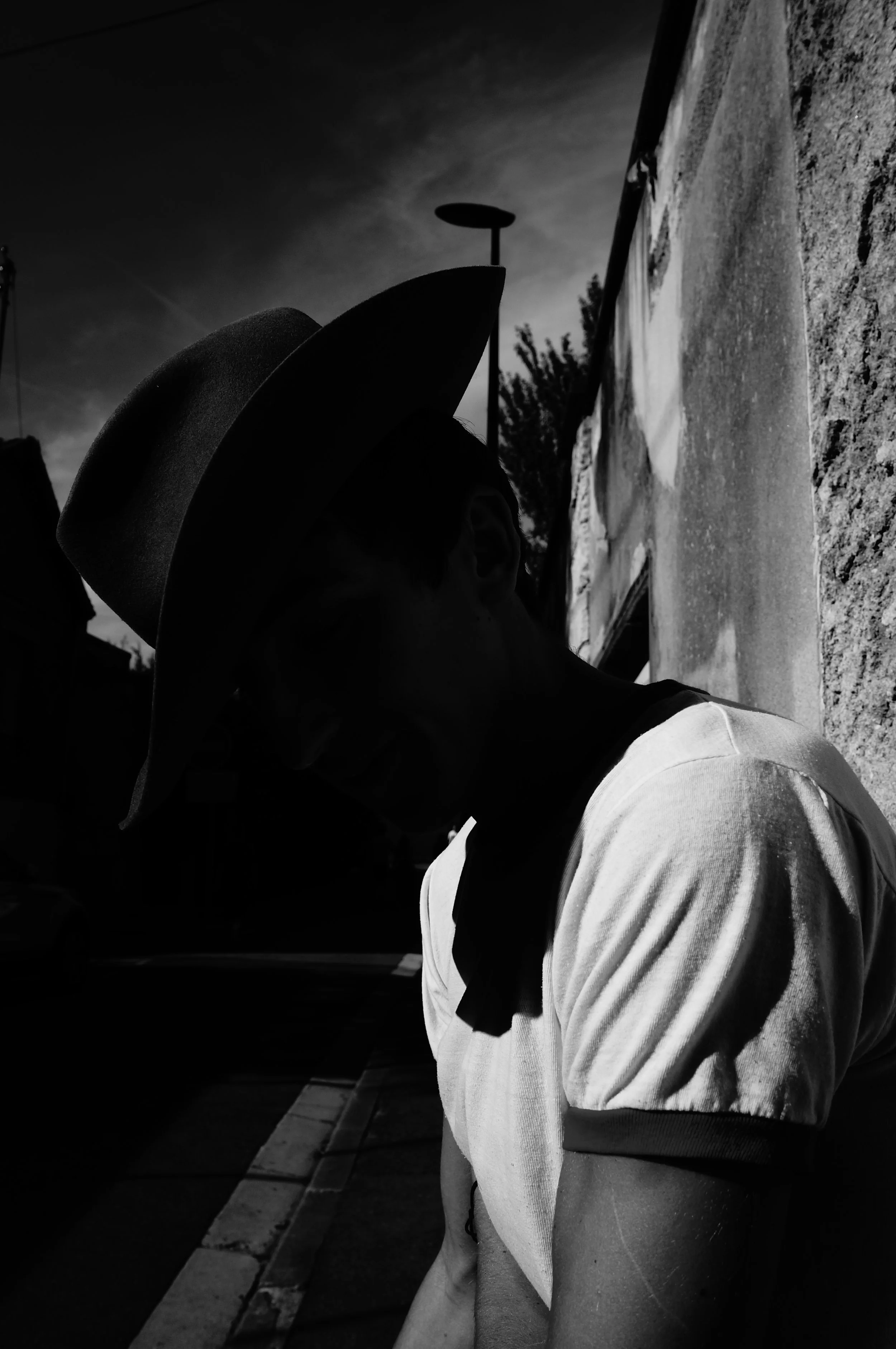 A silhouette of a person wearing a large hat, leaning against a textured wall on a street with a cloudy sky in the background.