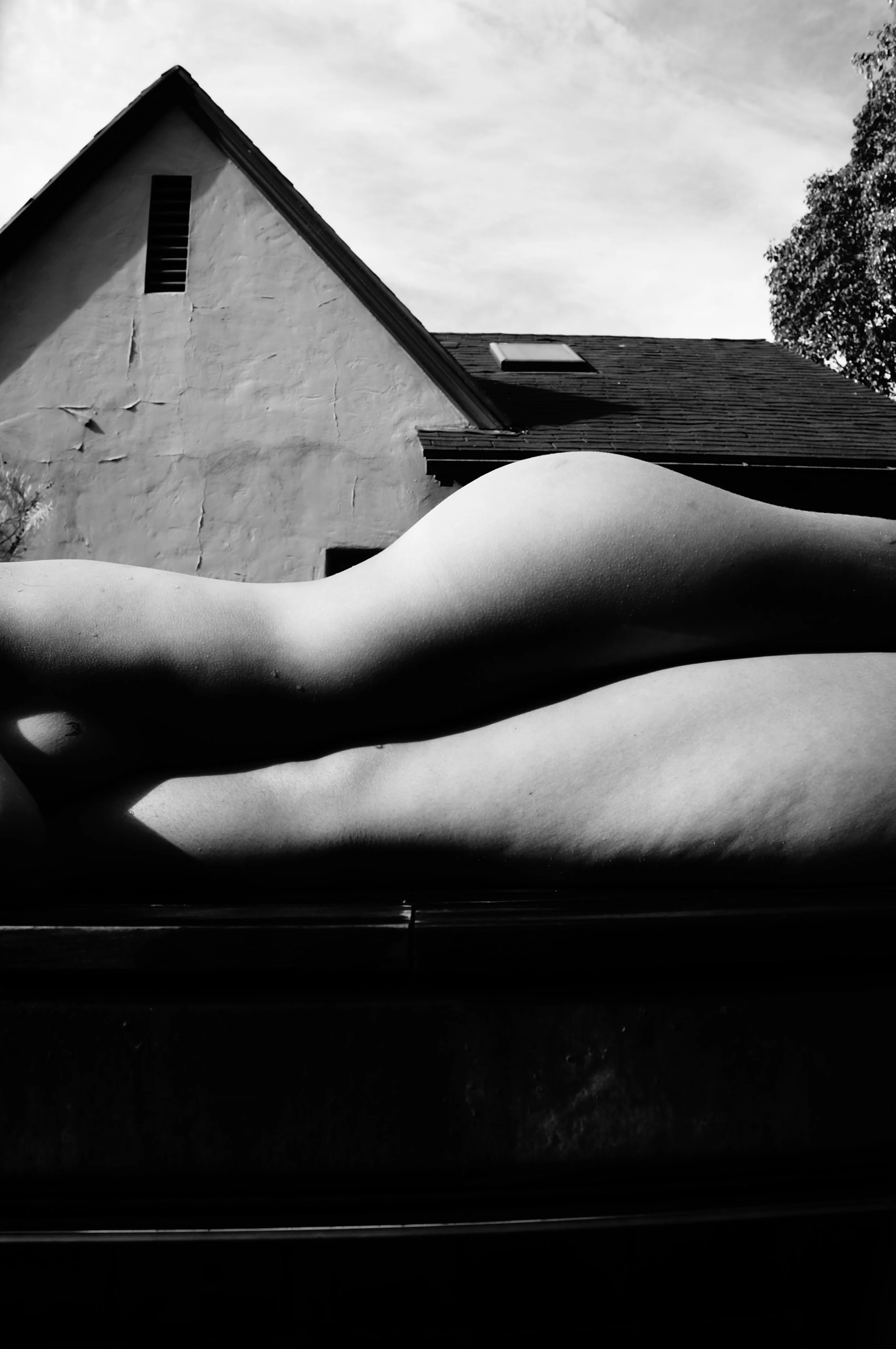 Black and white photo of a reclining nude figure with an arching back, lying on a dark surface outside, with a house and tree in the background.