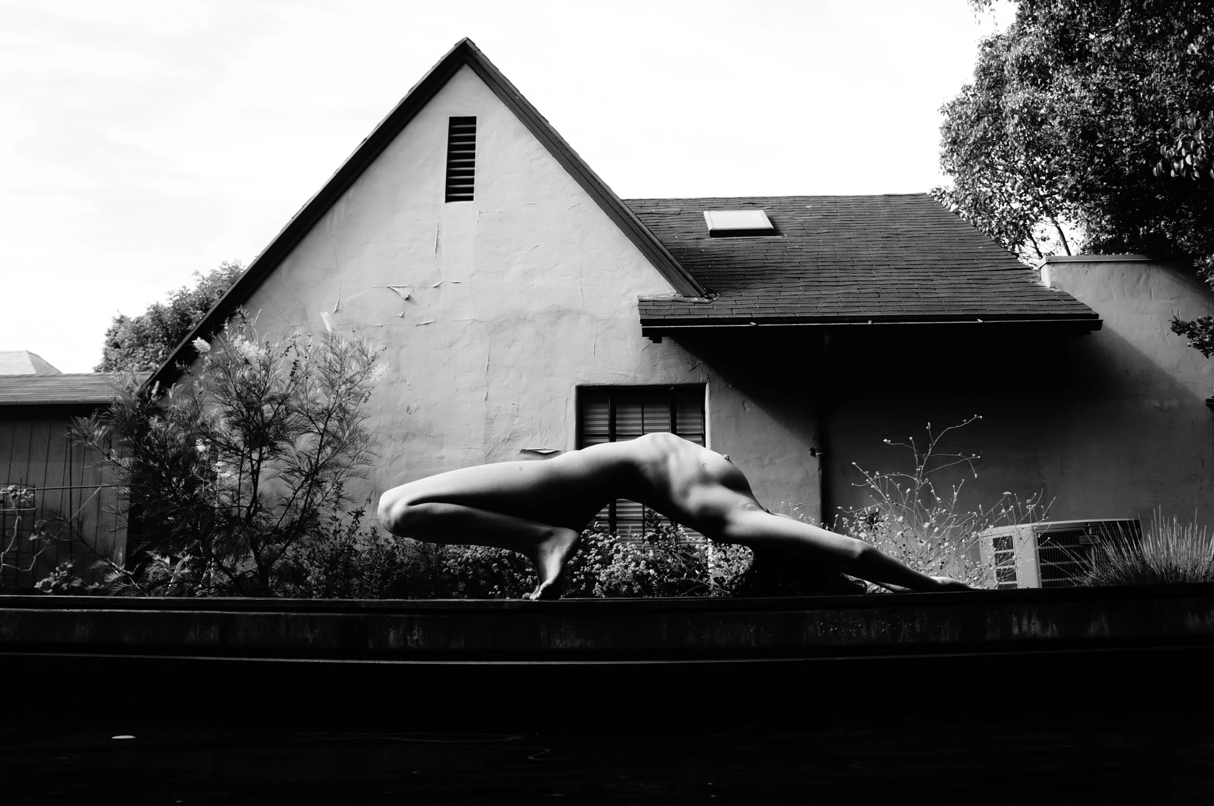 A nude person performing a yoga pose on a flat surface outside a house with a sloped roof and windows, surrounded by plants and trees in black and white.