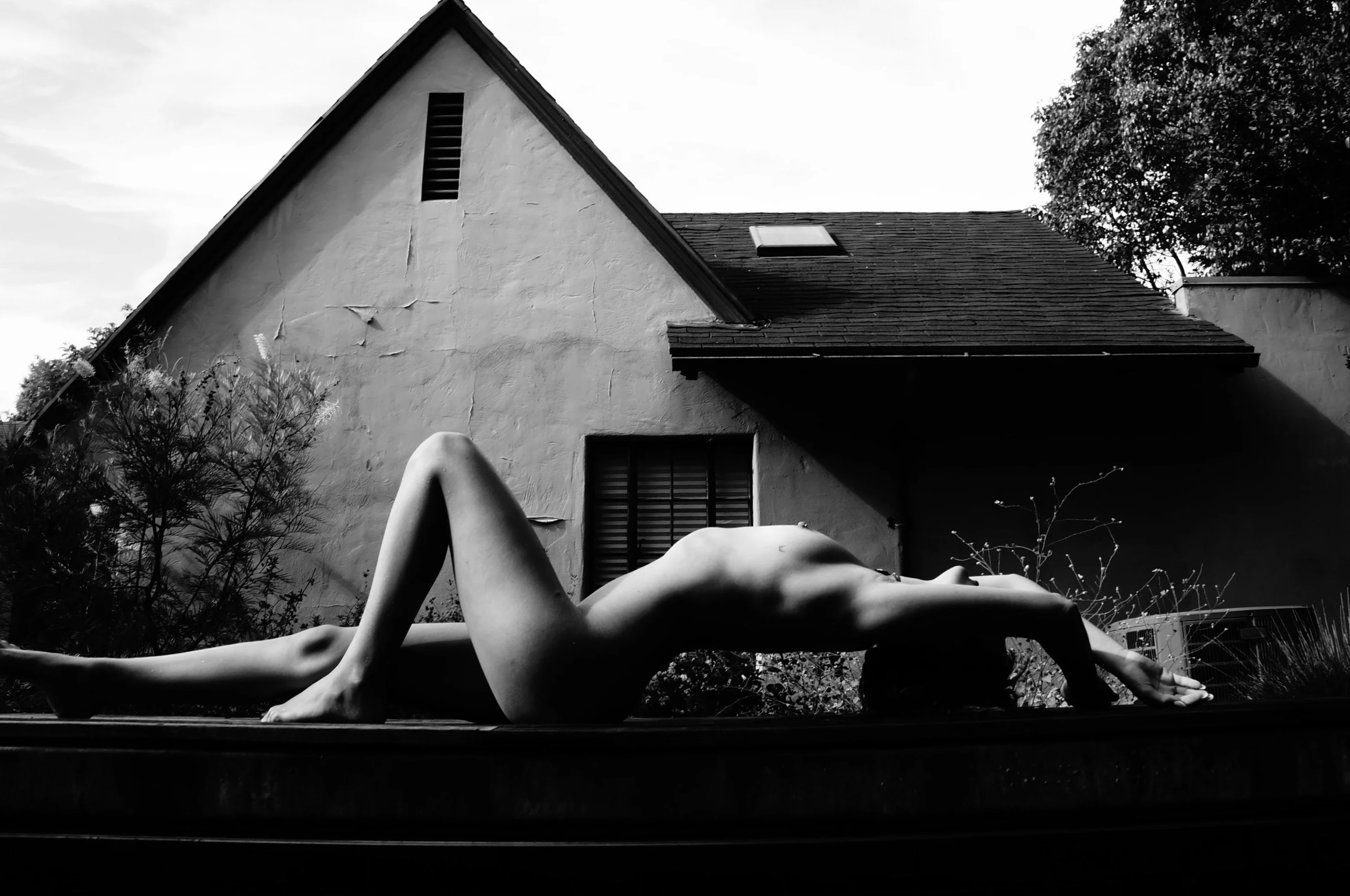 Black and white photo of a nude woman lying on her back on a surface outdoors, with her arms stretched above her head and her legs bent, in front of a house with a sloped roof and visible windows. There are trees and plants around the house.