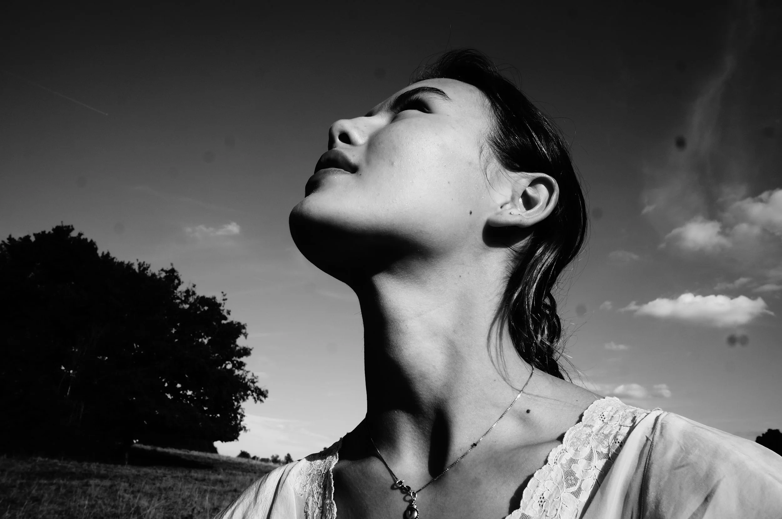 A black and white photo of a woman outdoors with her head tilted back, eyes closed, and face lifted towards the sky, with trees and a partly cloudy sky in the background.