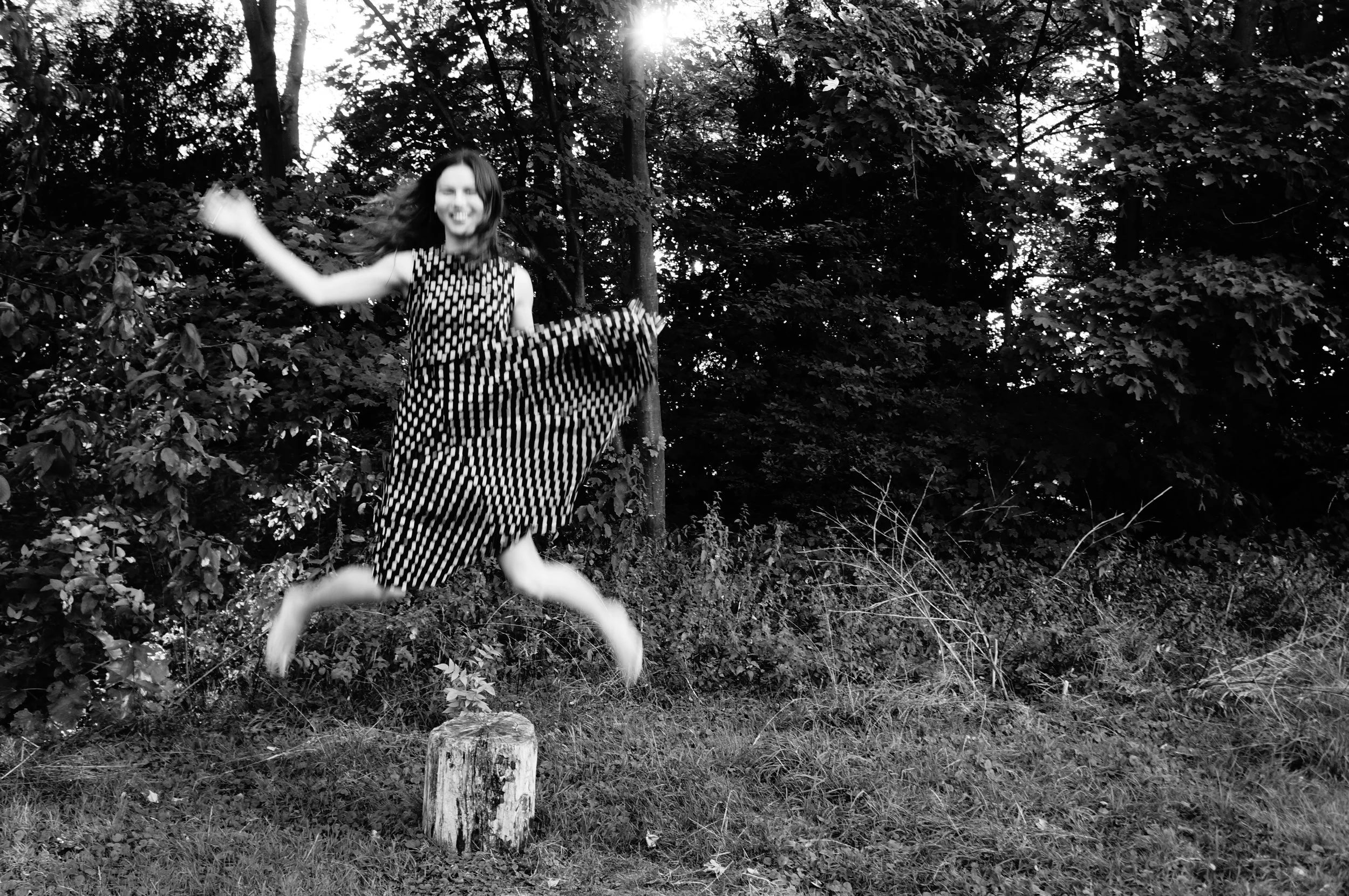 A woman in a polka dot dress jumping on a tree stump in a wooded area, smiling, in black and white.