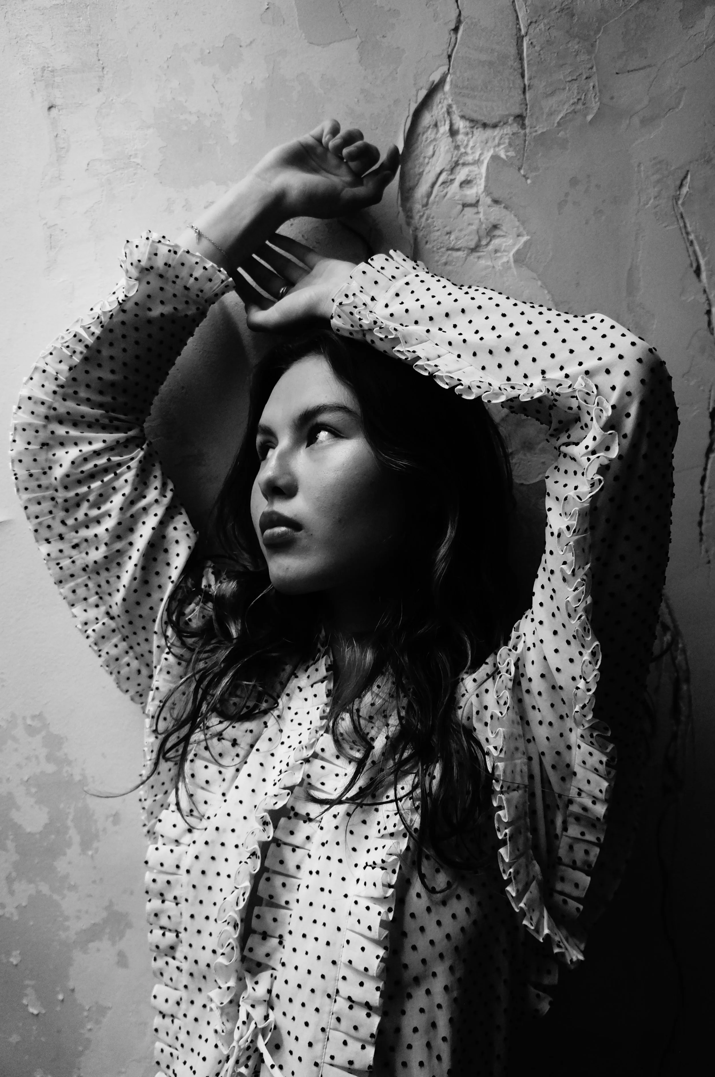 Black and white photo of a woman with long wavy hair, looking to the side with arms raised above her head, leaning against a textured wall.