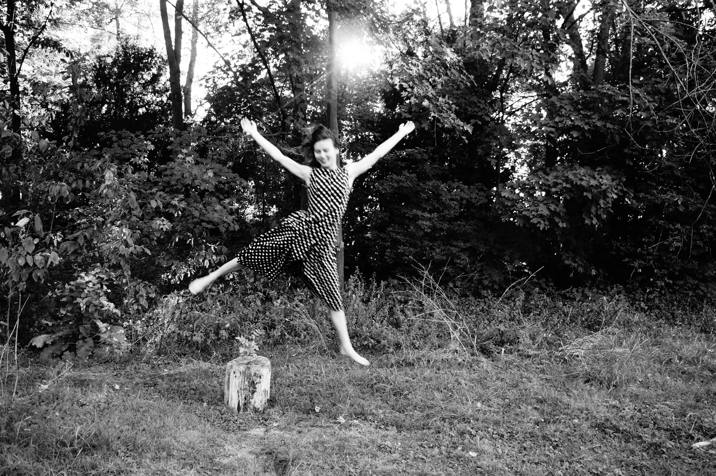 Black and white photo of a young girl in a polka dot dress jumping and smiling outdoors in a wooded area.