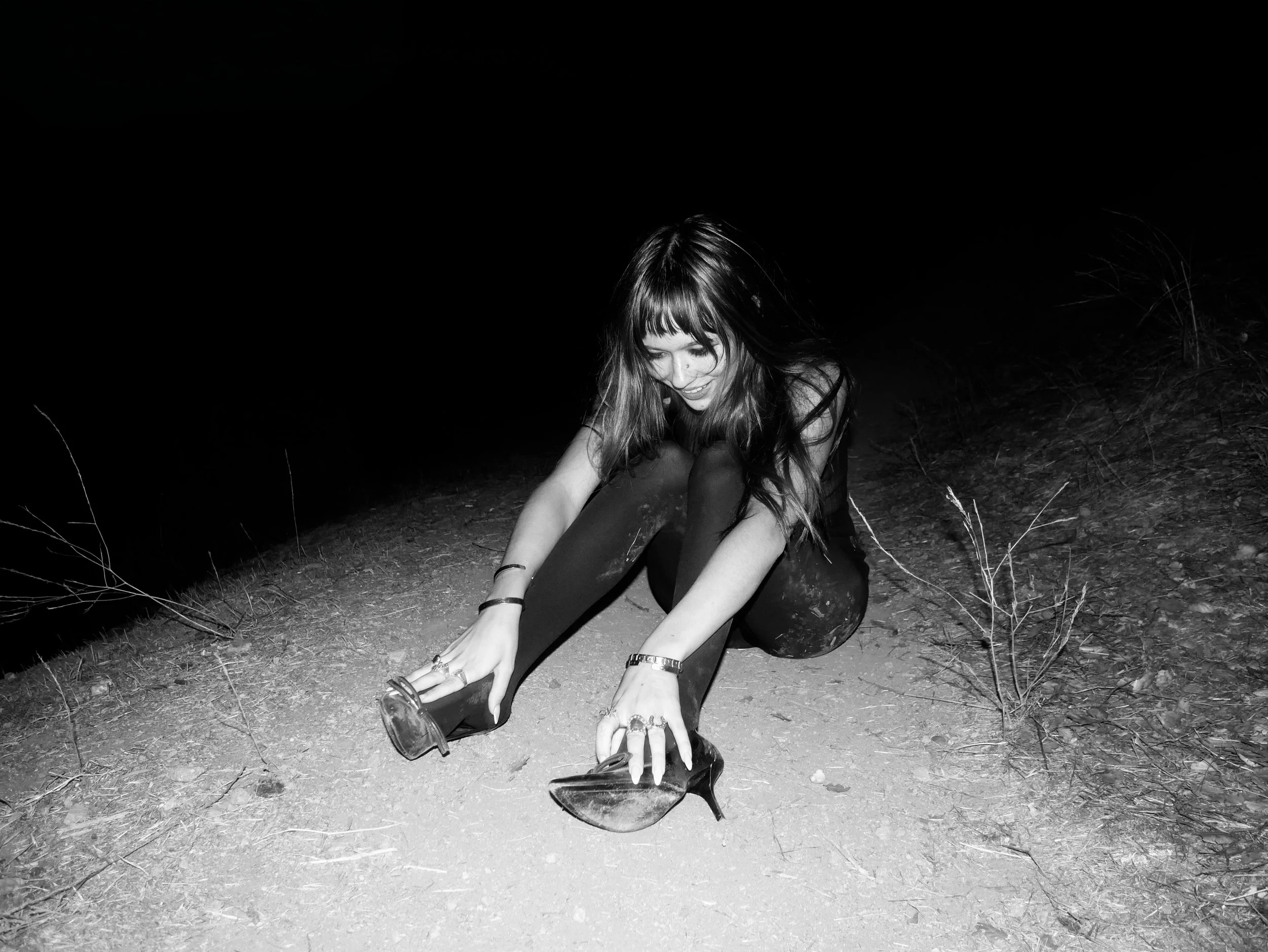 A woman with long hair and bangs, dressed in dark clothing, is sitting on the ground outdoors at night, putting on high-heeled shoes, with a smile on her face.